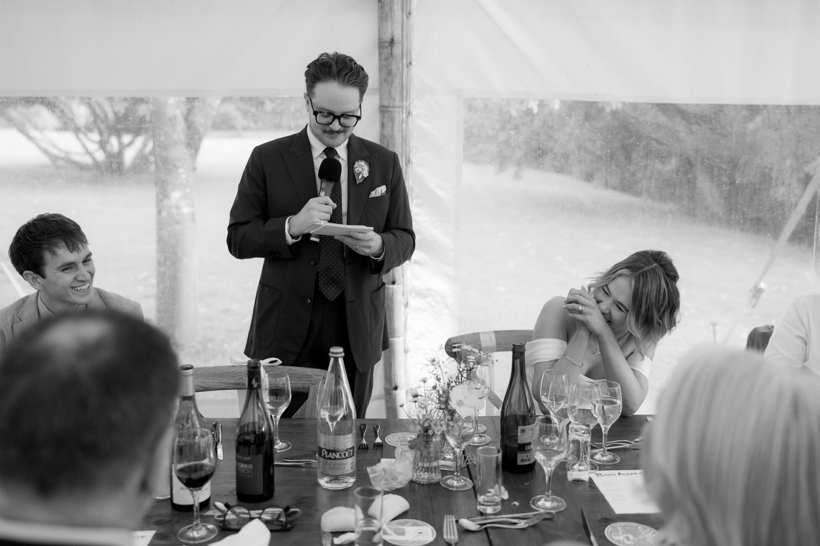 A man in a suit stands and speaks from a note at a table set for a meal; people around him are seated, with one woman laughing and covering her face.