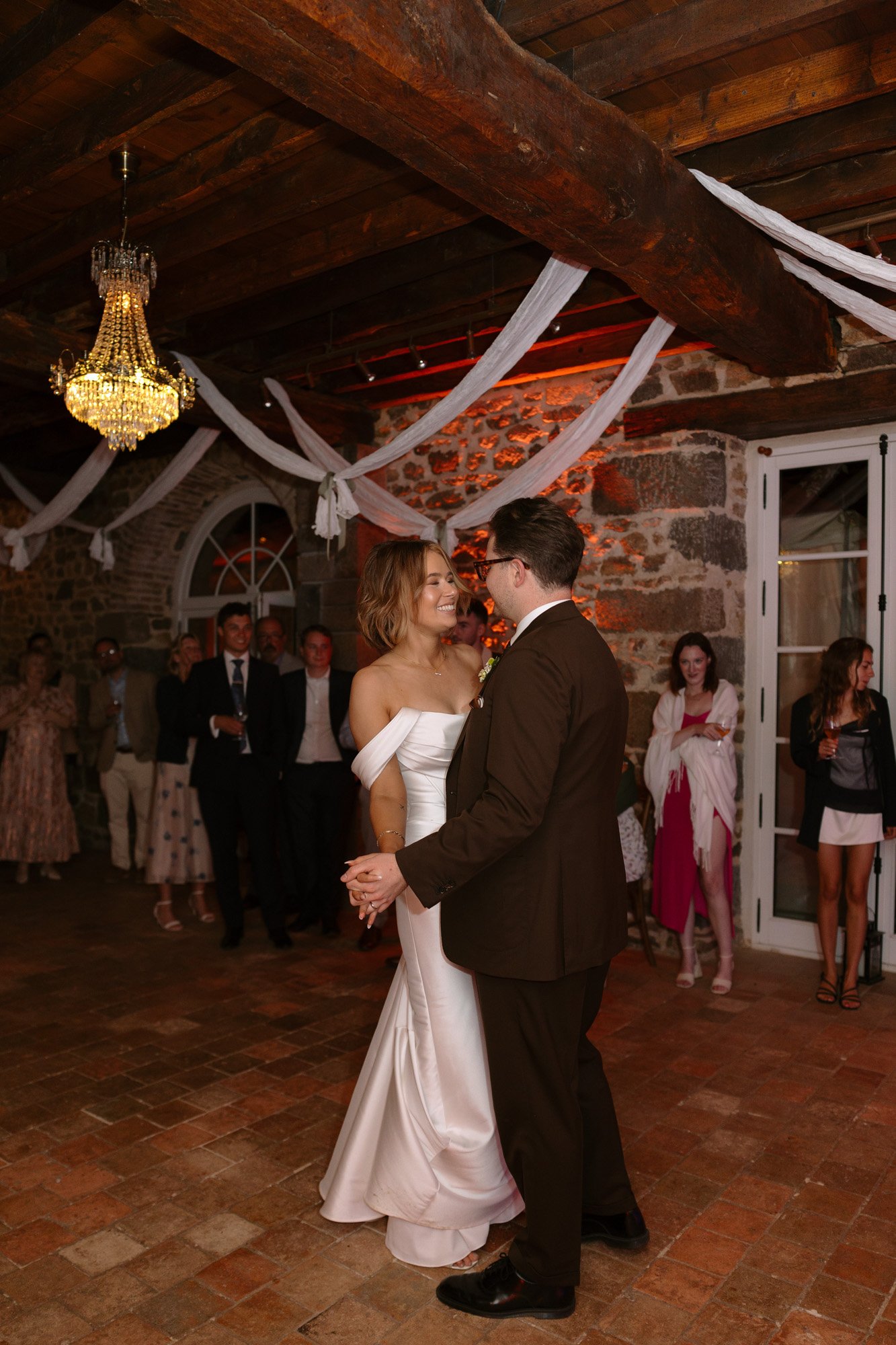 A bride and groom dance together in a rustic indoor venue with stone walls and wooden beams, while guests watch in the background.