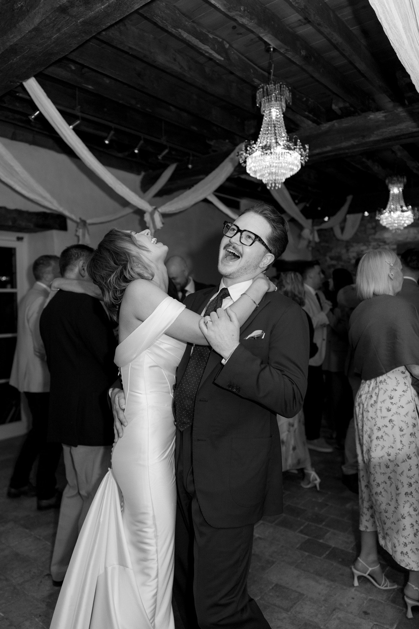 A woman in a white dress and a man in a suit laugh and dance together at an indoor event with chandeliers and other guests in the background.