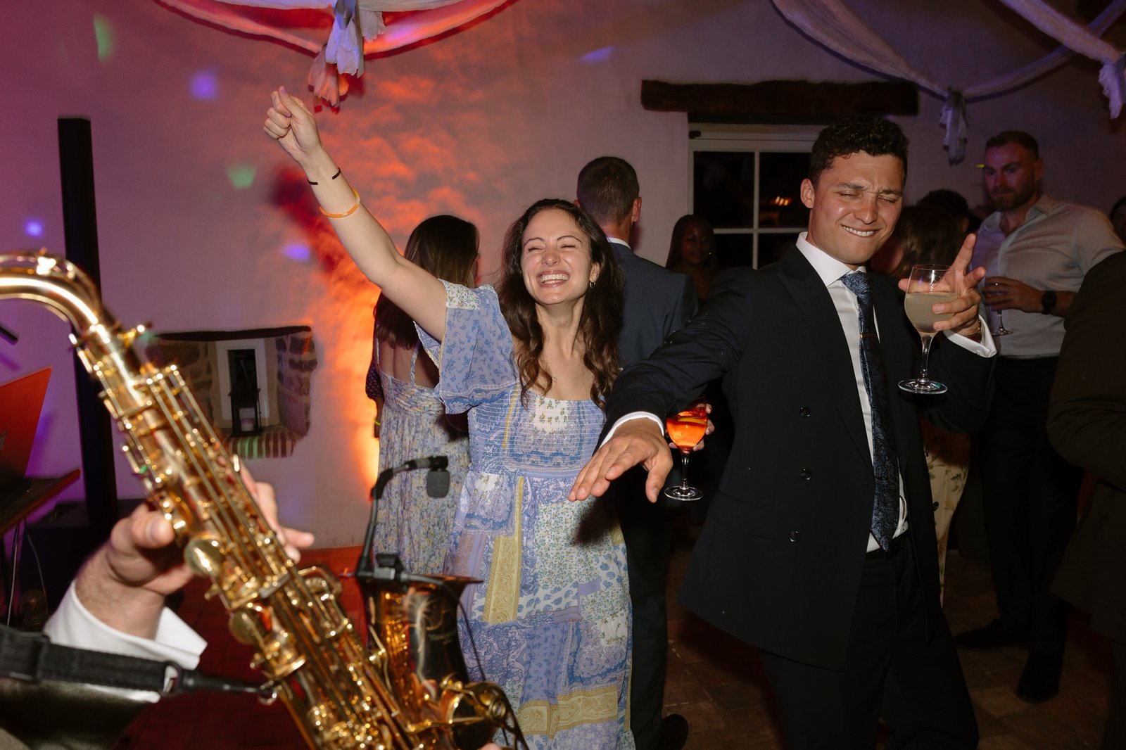 A woman and a man dance and smile at a party, holding drinks, while a saxophonist plays in the foreground. Other guests are visible in the background.
