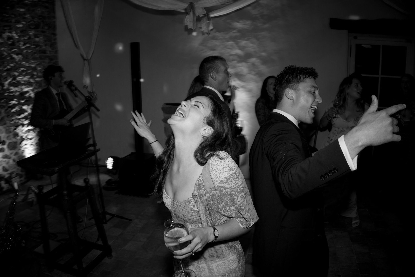 A woman and man in formal attire dance and laugh at an indoor event, with other guests and a musician visible in the background.