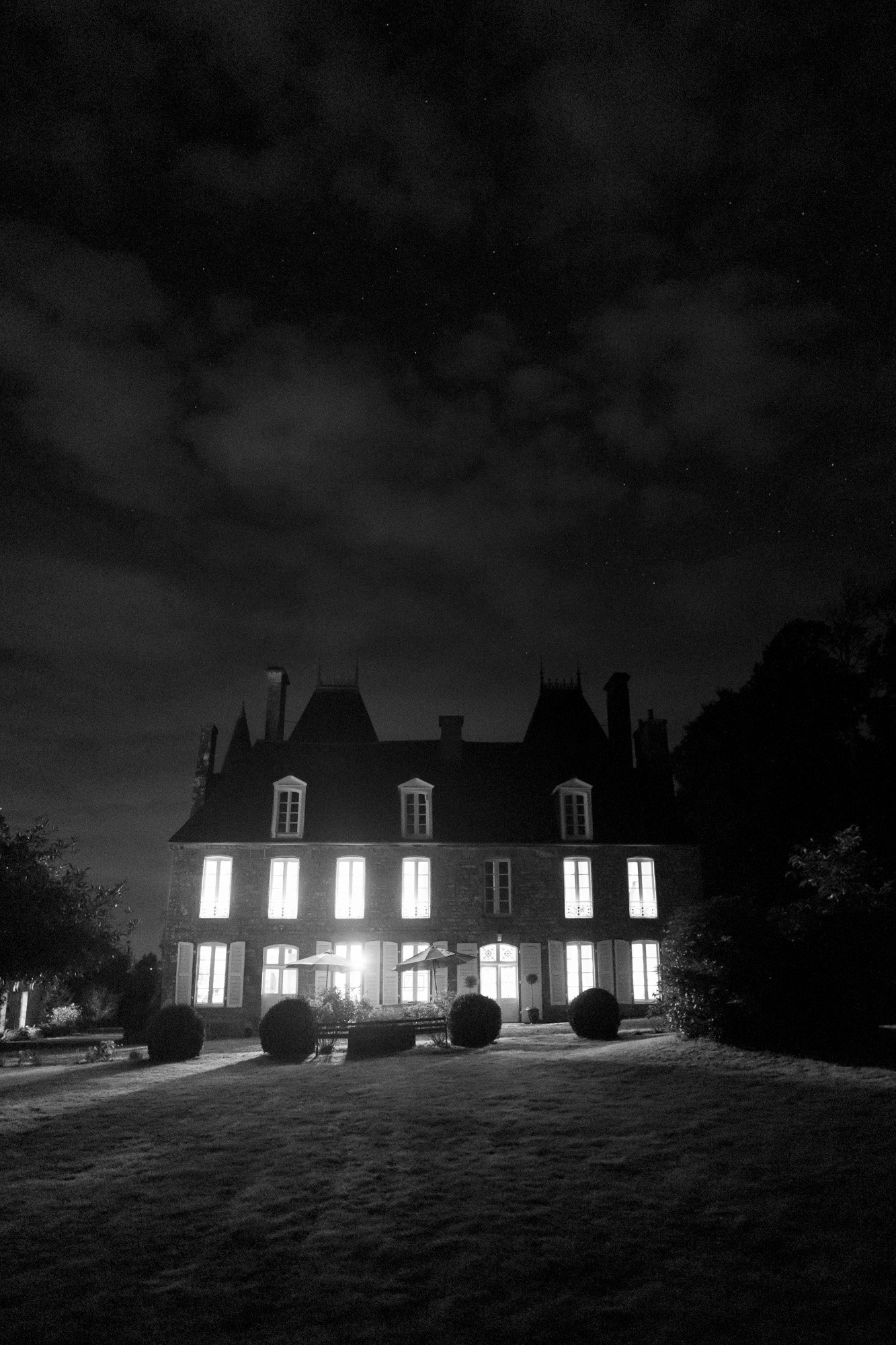 A large, dark mansion with illuminated windows stands under a cloudy night sky, surrounded by shadows and trees.