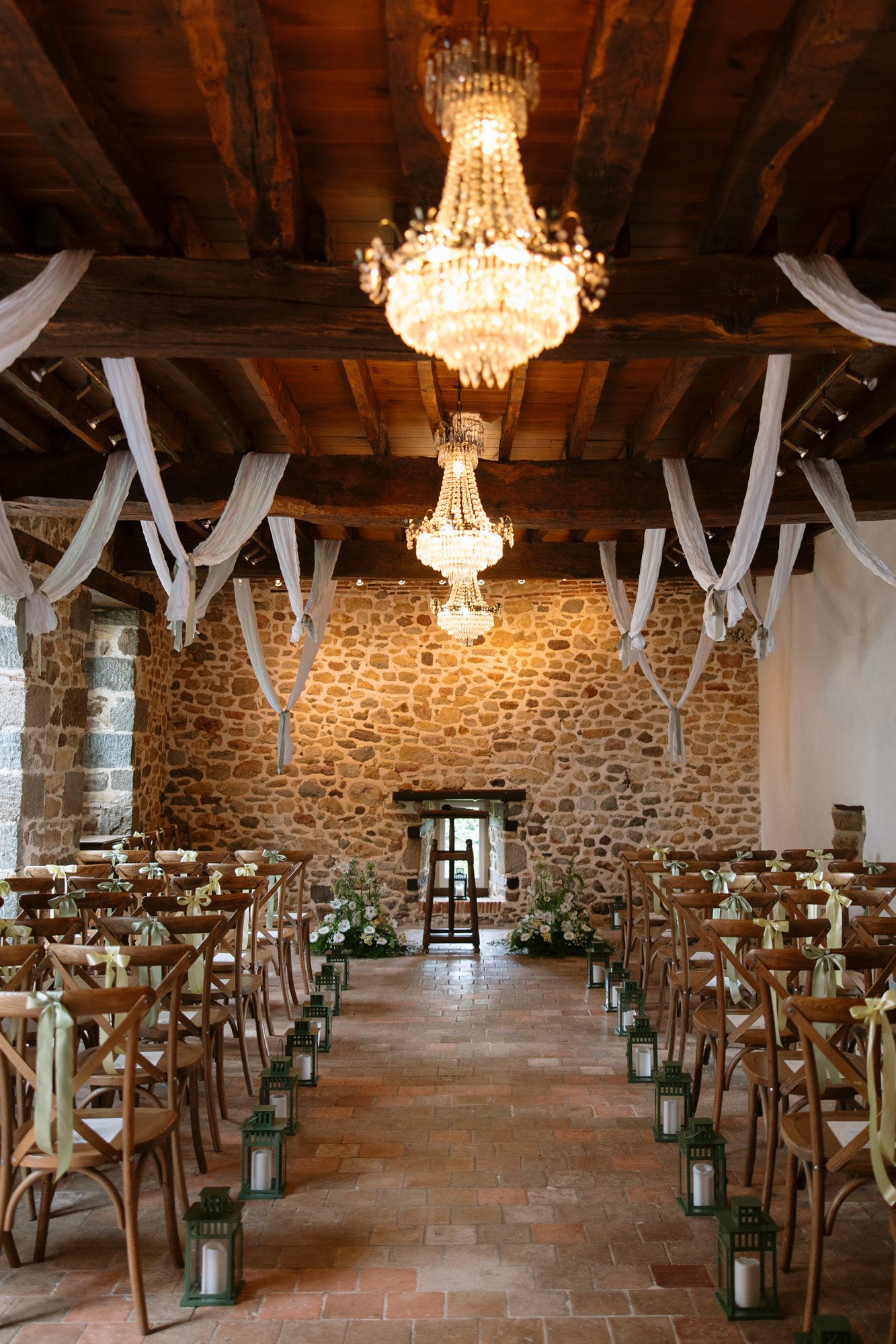 A rustic indoor wedding ceremony setup with wooden chairs, chandeliers, draped white fabric, stone walls, and lanterns lining the aisle.