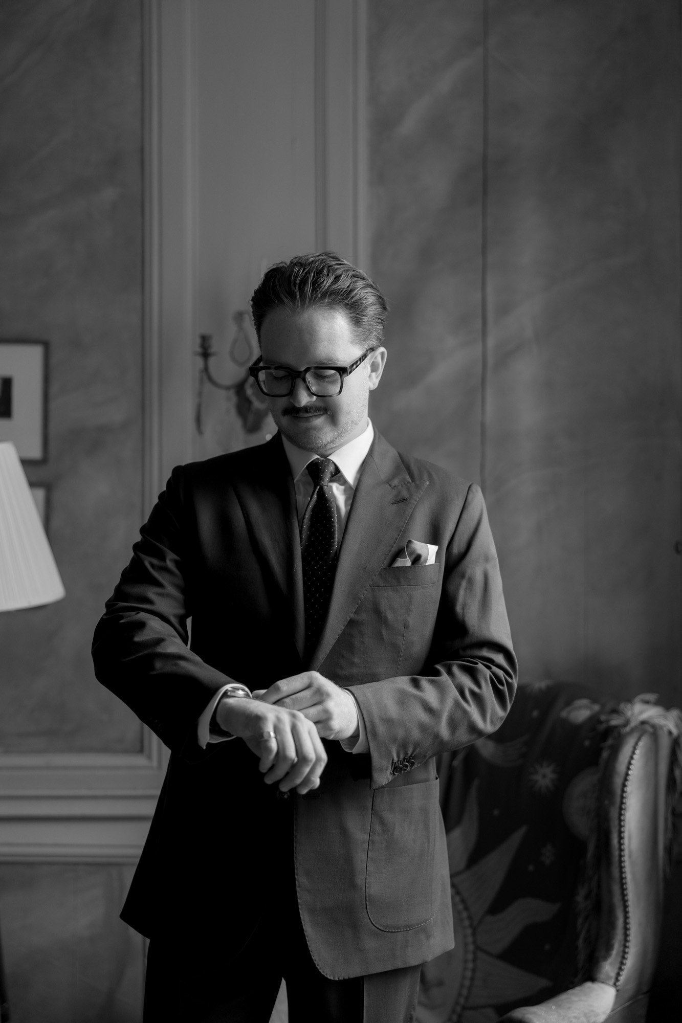 A man in a suit and glasses stands indoors, looking at his wristwatch.