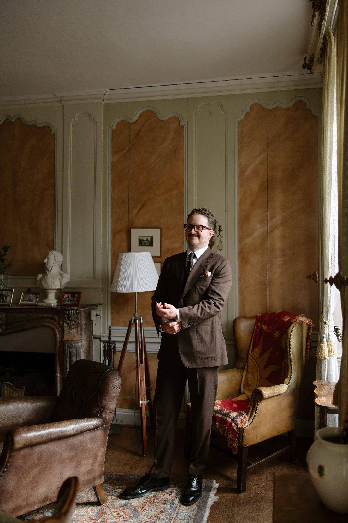 A man in a brown suit stands in a traditionally decorated living room with wooden paneling, vintage furniture, and a patterned rug.