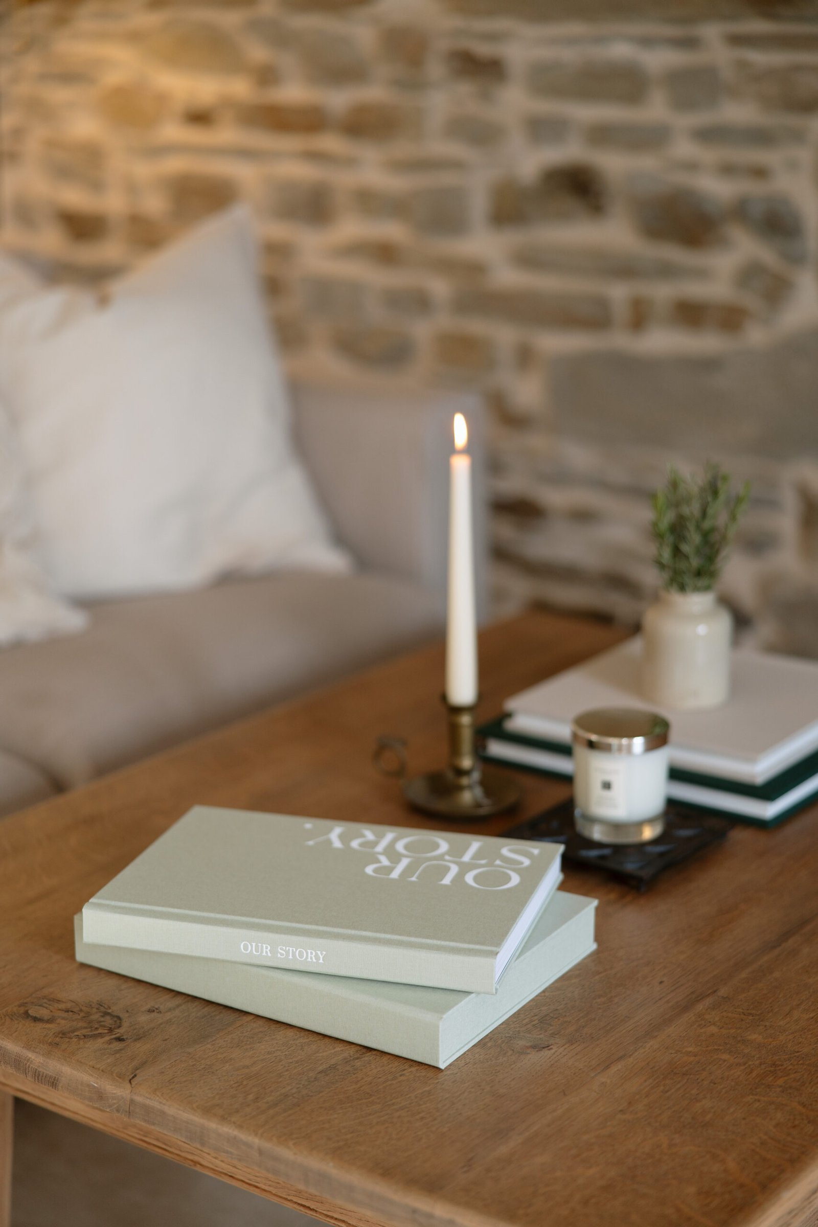 A wooden table with a book titled Our Story, a lit white candle in a brass holder, a small potted plant, and a candle jar, with a sofa and stone wall in the background.