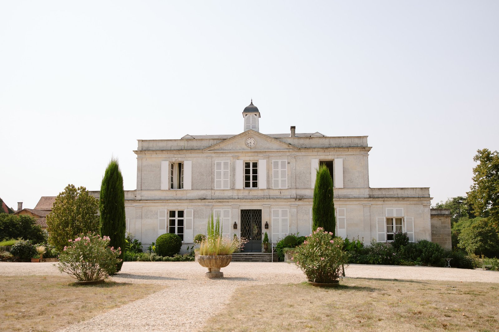A large, symmetrical stone building with white shutters, a central clock, tall windows, and manicured landscaping in front, set under a clear sky.  Captured by Cognac wedding photographer.