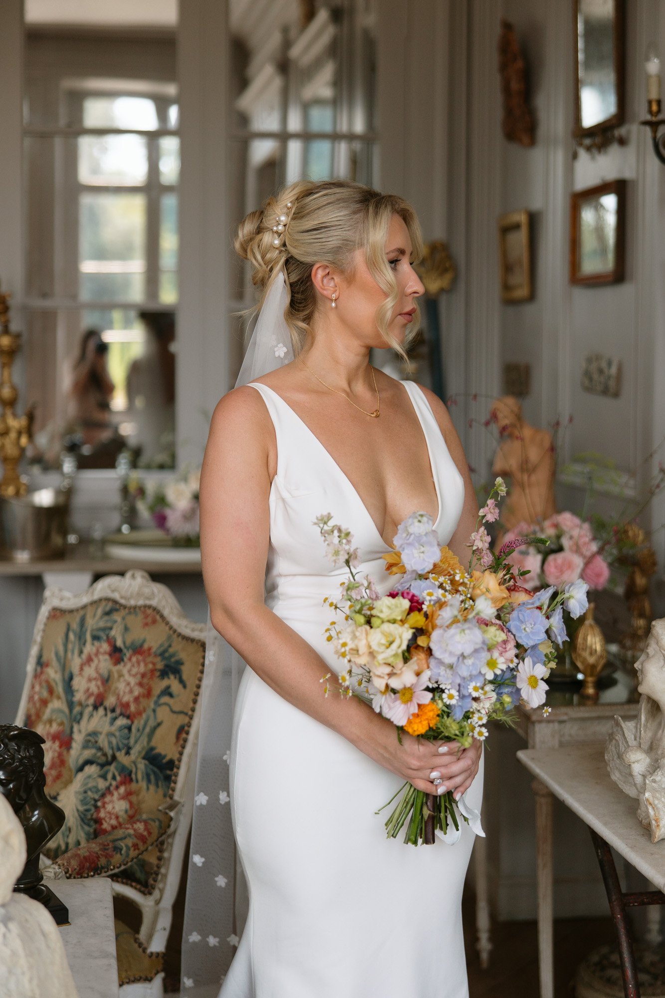 A woman in a white sleeveless wedding dress holds a colorful bouquet in an ornately decorated room, looking to the side. Captured by Cognac wedding photographer.