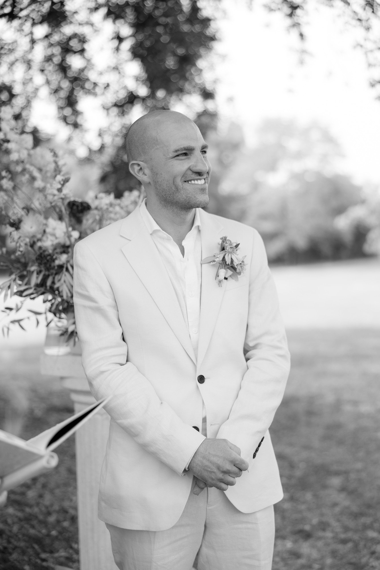 A man in a light-colored suit stands outdoors, smiling, with hands clasped. He wears a boutonniere and trees and flowers are visible in the background.