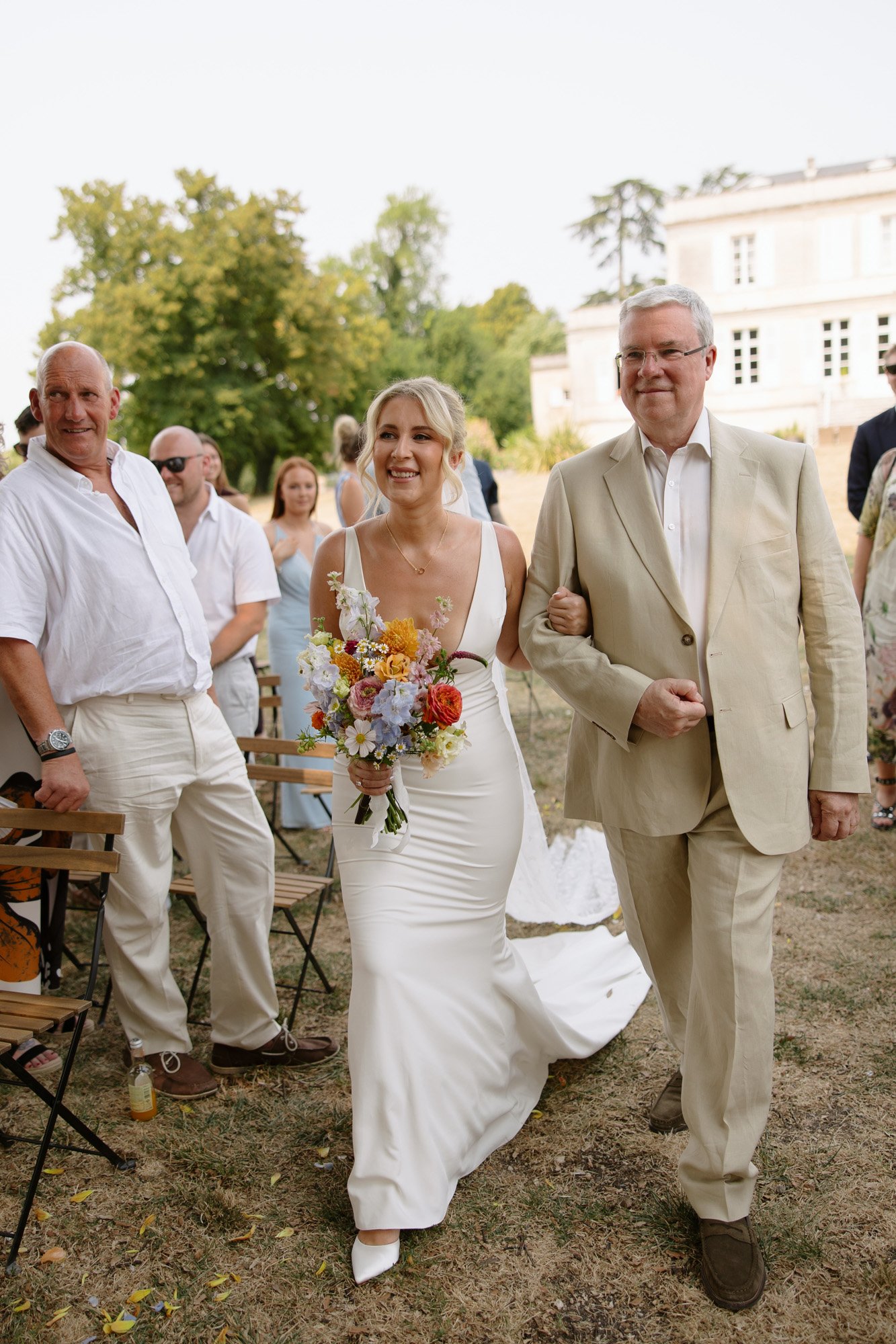 A bride in a white dress walks down the aisle outdoors, holding a bouquet and escorted by an older man in a beige suit, while guests look on.