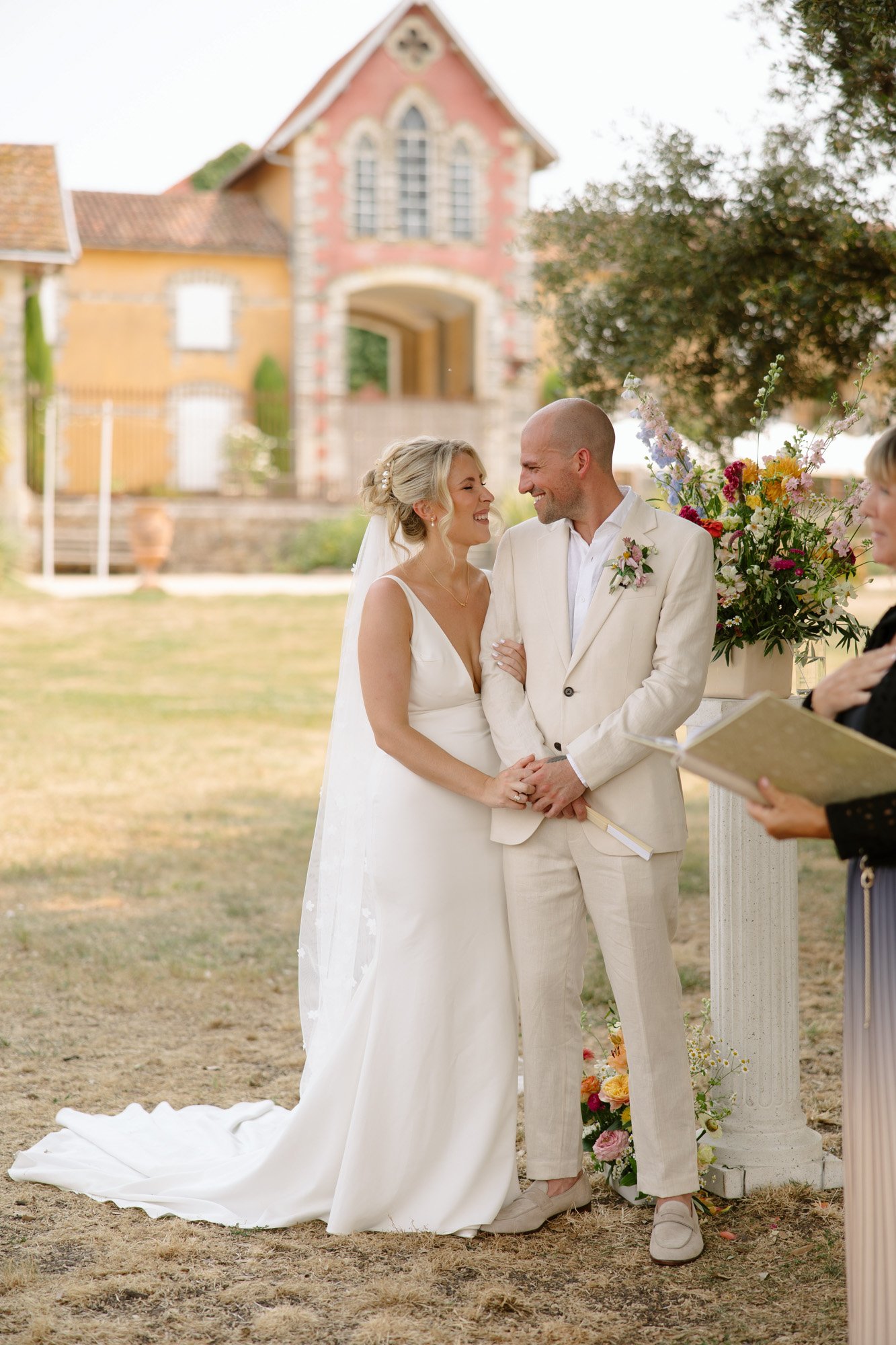 A bride and groom stand together outdoors, smiling at each other in front of a flower arrangement, with a building in the background. Captured by Cognac wedding photographer.