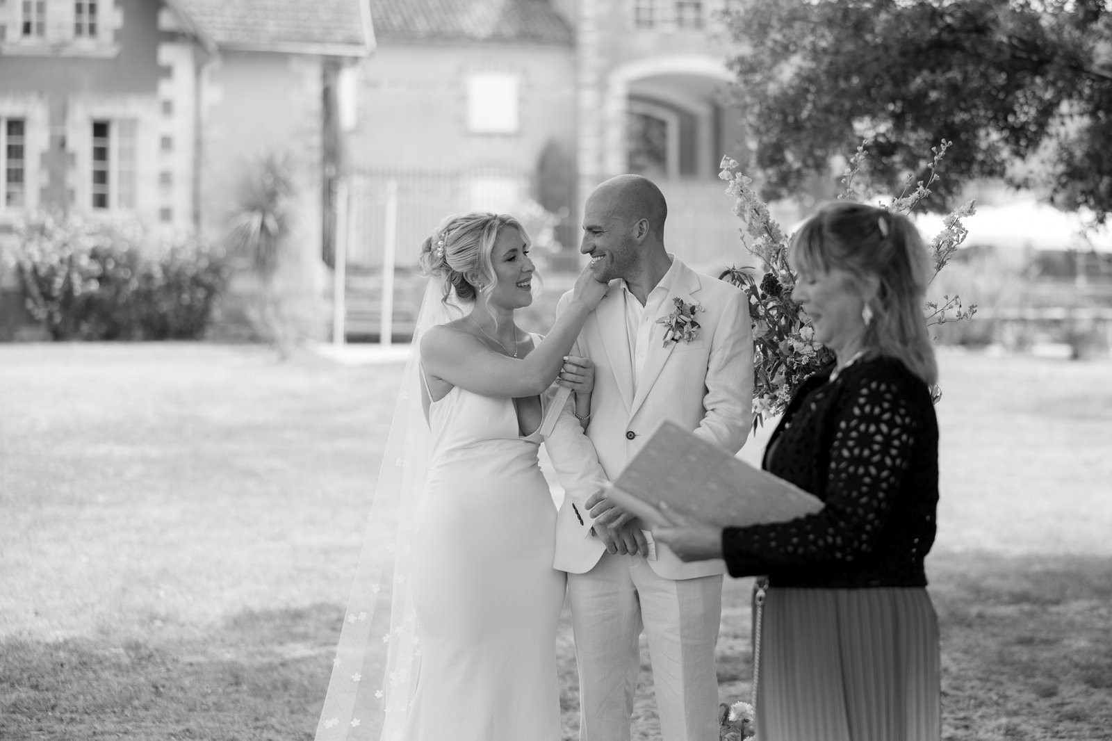 A bride and groom stand together smiling during an outdoor wedding ceremony, while an officiant holds a book in the foreground. Captured by Cognac wedding photographer.