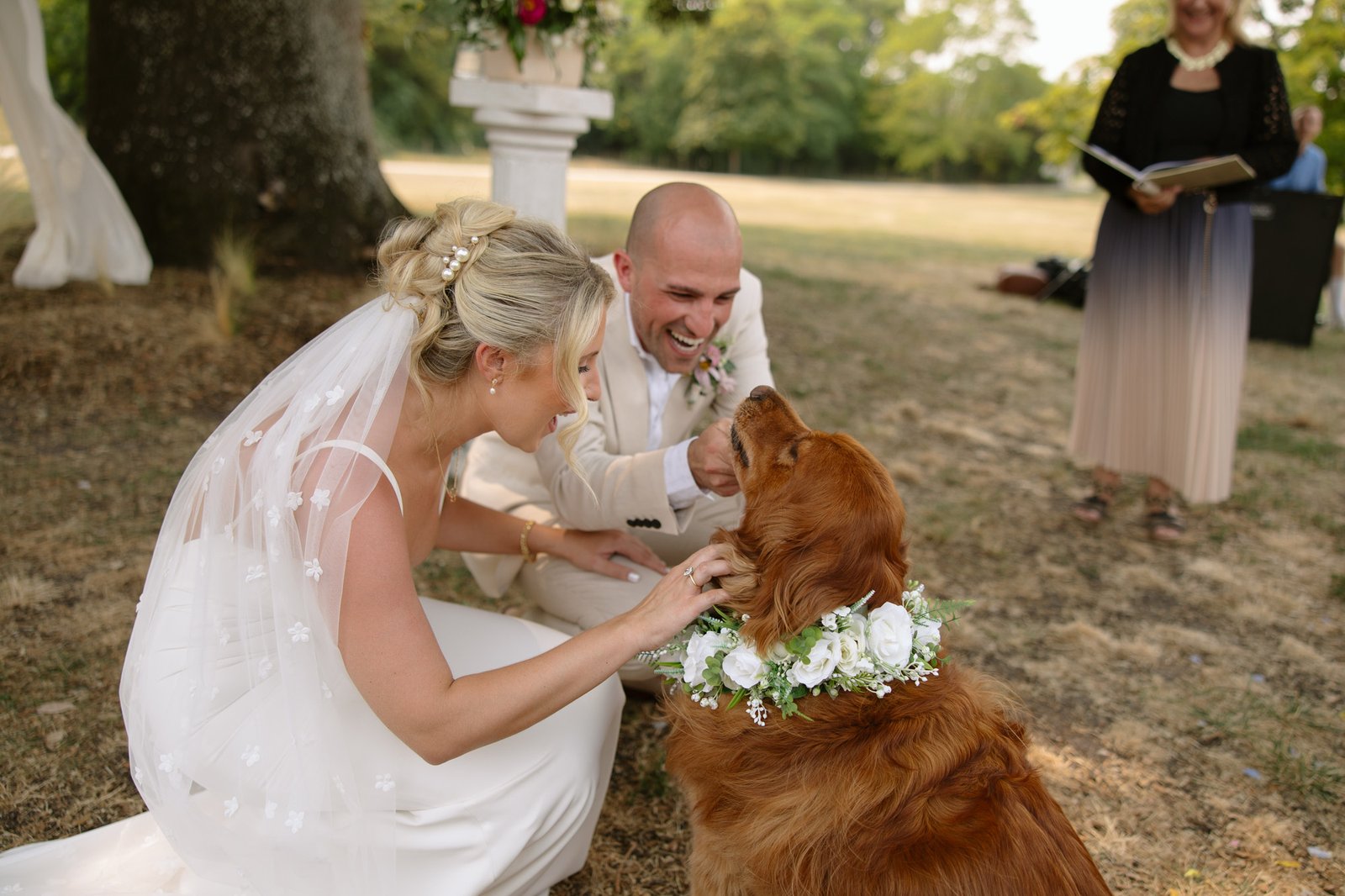A bride and groom kneel outdoors, smiling and petting a golden retriever wearing a white floral collar.