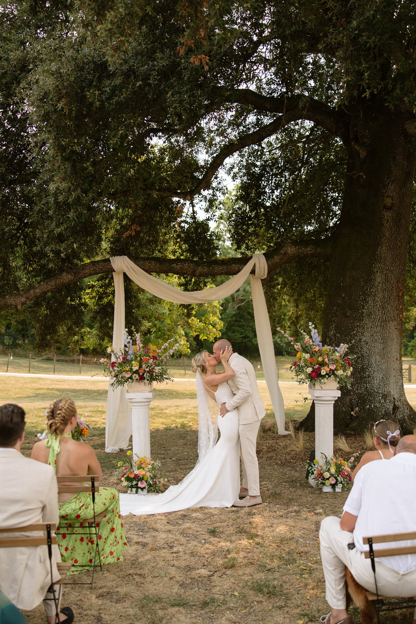 A bride and groom kiss at an outdoor wedding ceremony under a large tree, surrounded by guests seated on chairs.