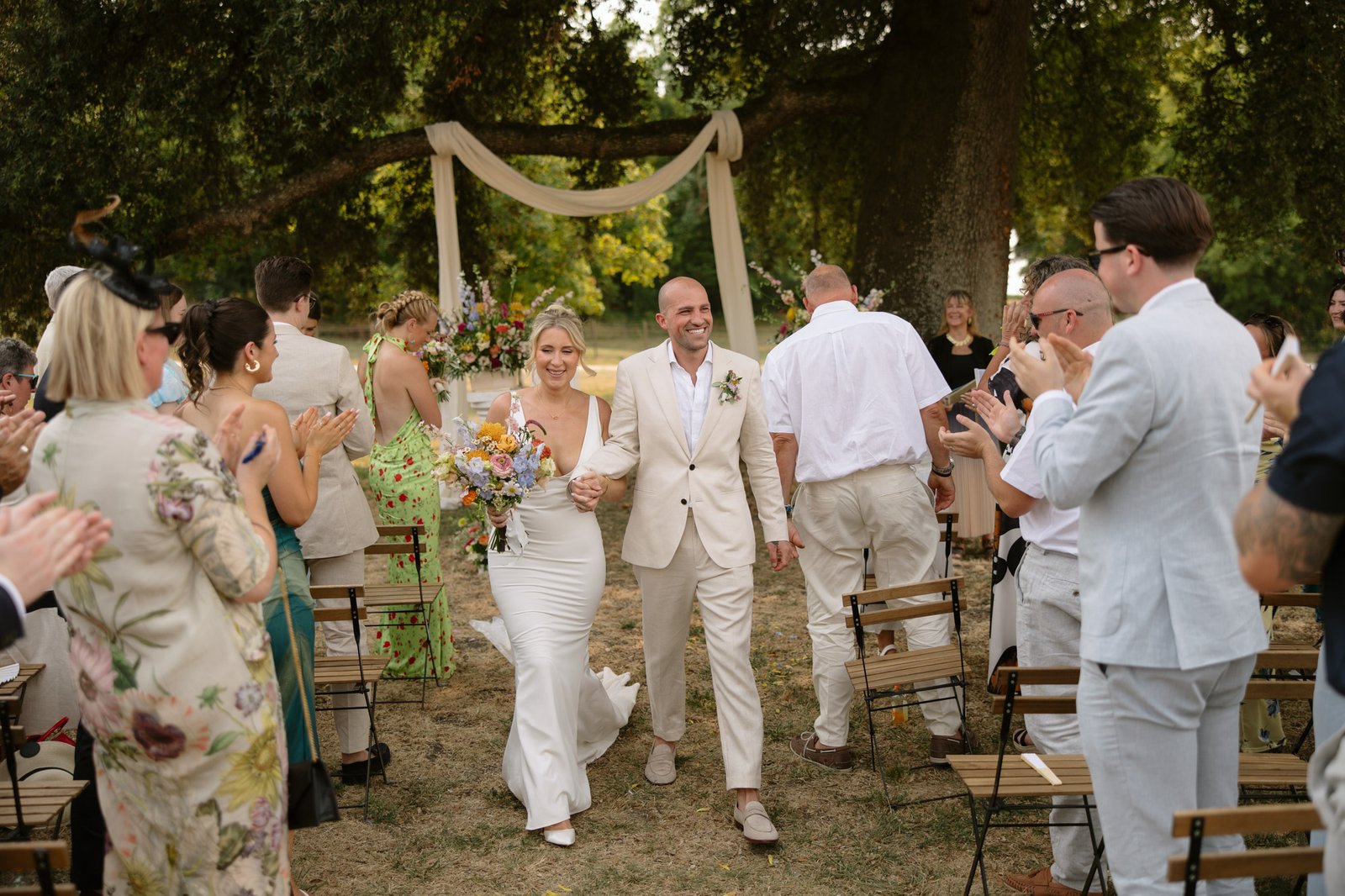 A bride and groom walk down the aisle outdoors, smiling and holding hands, as guests stand and clap.