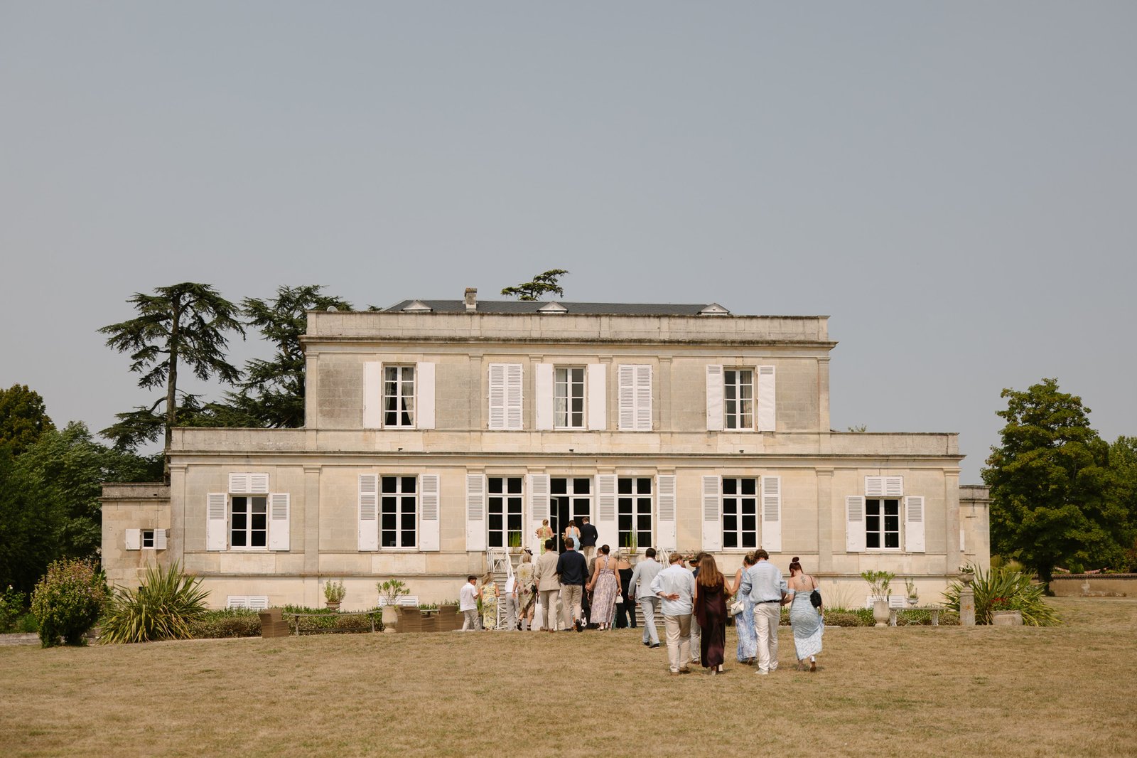 A group of people stands outside and enters a large, two-story beige building with many windows, surrounded by grass and trees on a clear day.