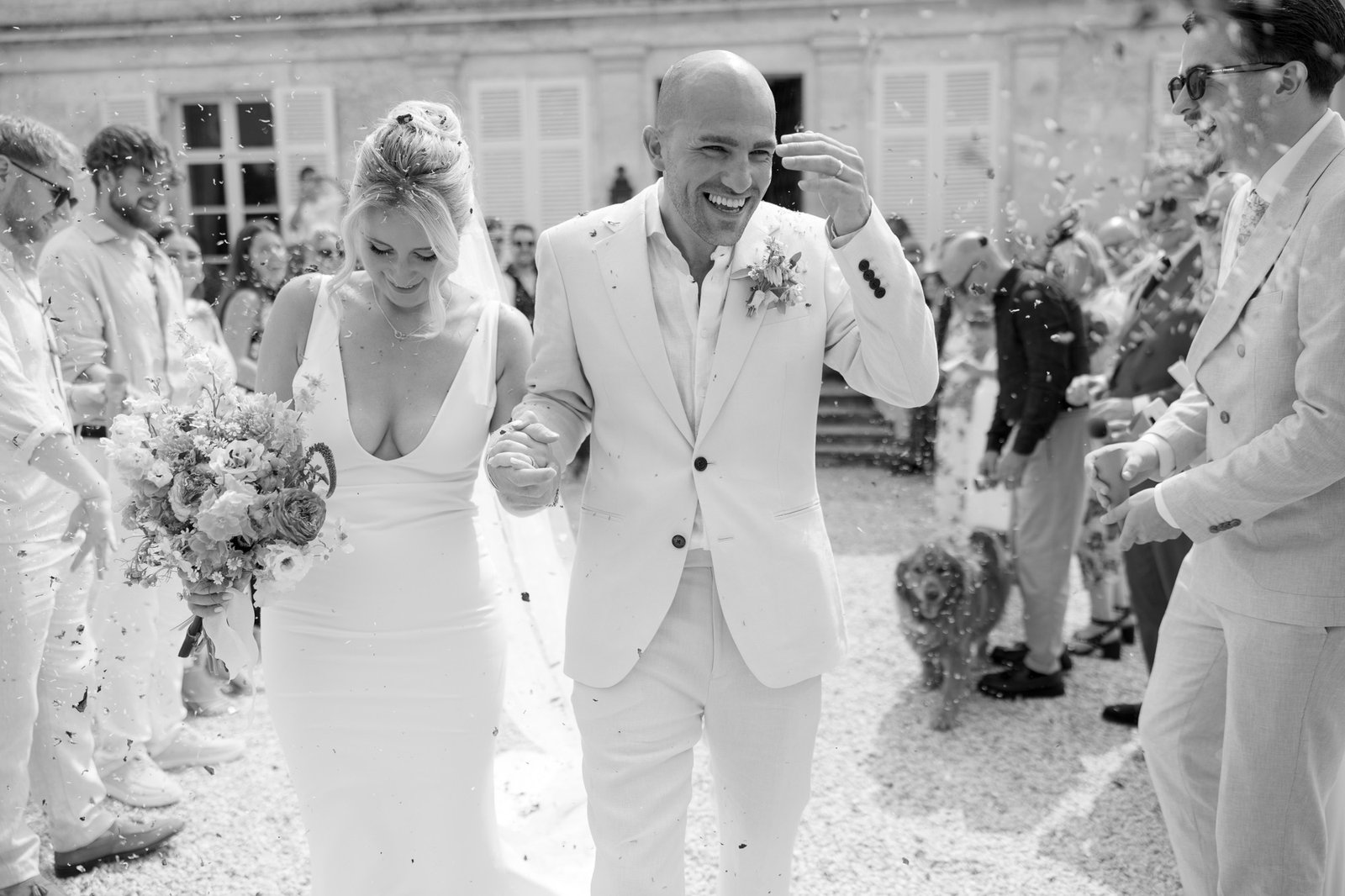A bride and groom walk outdoors, smiling and holding hands, surrounded by guests tossing confetti at their wedding. Captured by Cognac wedding photographer.