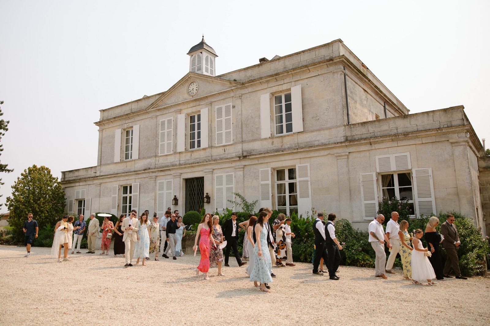 A group of people in formal attire gather outside a large, light-colored, historic building with tall windows and a clock on the roof. Captured by Cognac wedding photographer.
