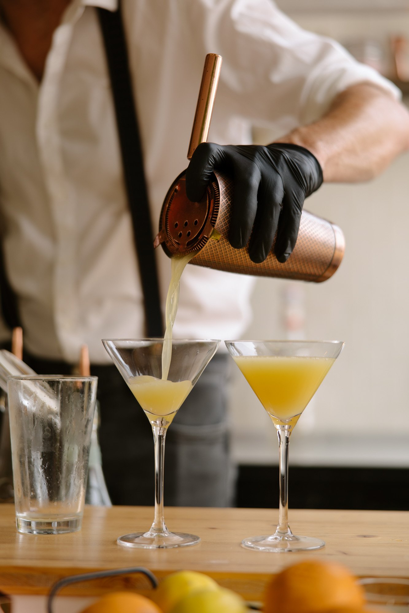 A person wearing black gloves pours a yellow cocktail from a shaker into two martini glasses on a wooden counter.