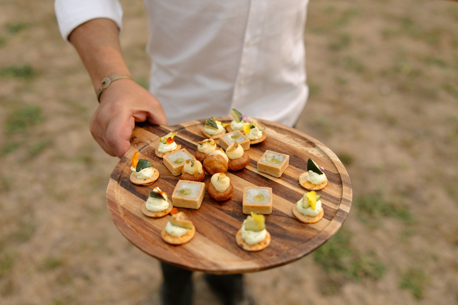A person in a white shirt holds a round wooden tray with assorted bite-sized appetizers, including canapés and pastries, displayed outdoors.