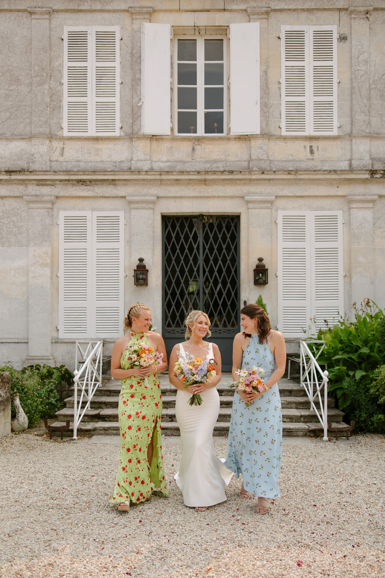 Three women in formal dresses, holding bouquets, stand in front of a stone building with white shutters and steps. Captured by Cognac wedding photographer. Captured by Cognac wedding photographer.