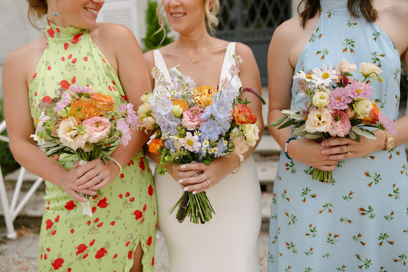 Three women in floral dresses and a white dress hold colorful bouquets of flowers, standing side by side outdoors.
