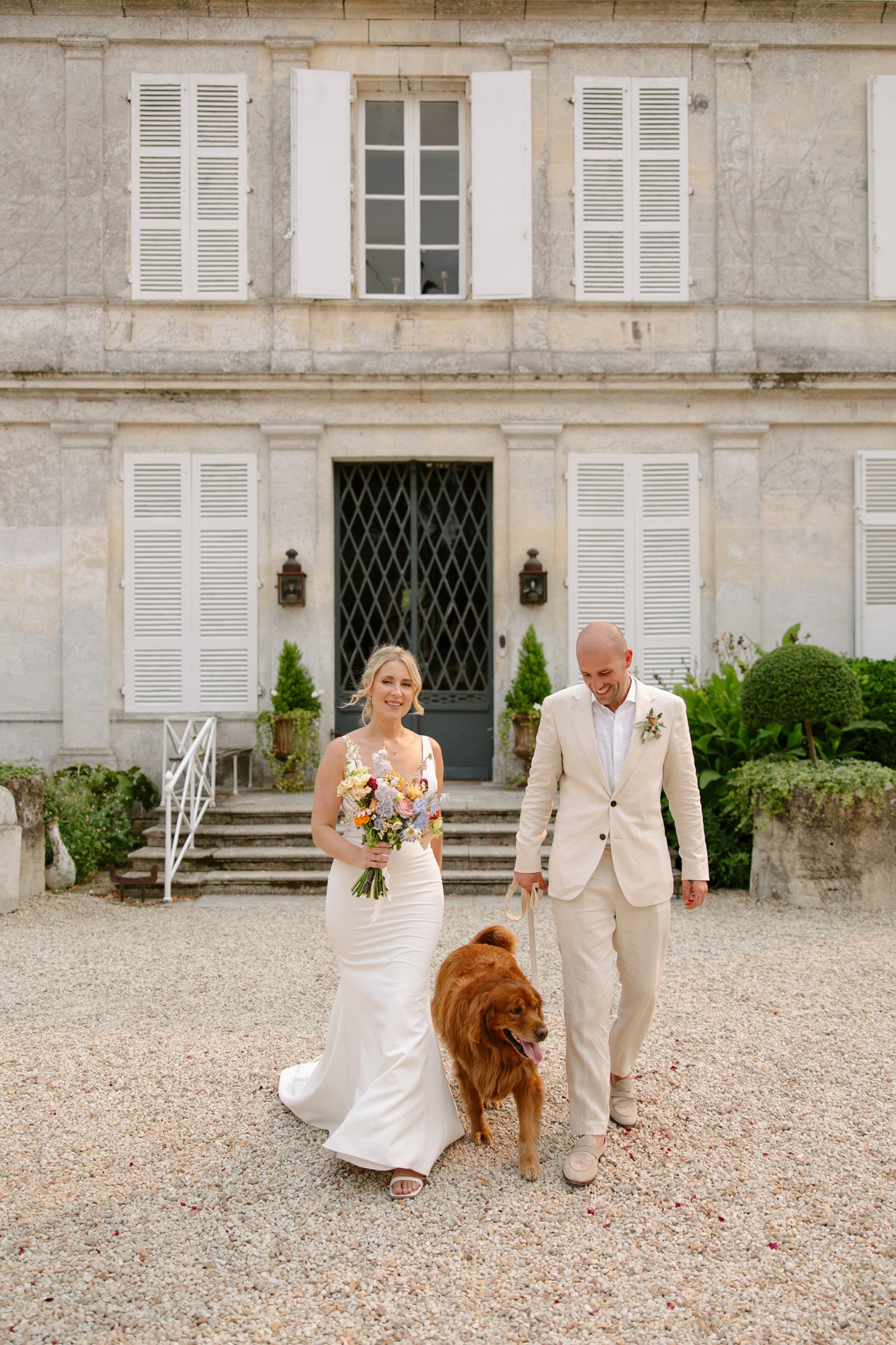 A bride and groom in cream-colored attire walk with a large brown dog in front of a white stone building with shuttered windows. Captured by Cognac wedding photographer.