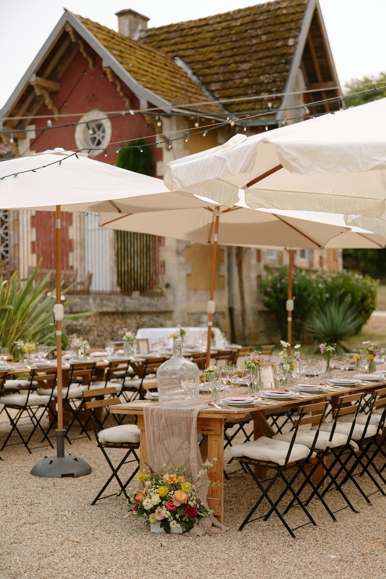 Outdoor dining setup with wooden tables, folding chairs, white umbrellas, floral arrangements, and a rustic building in the background. Captured by Cognac wedding photographer.