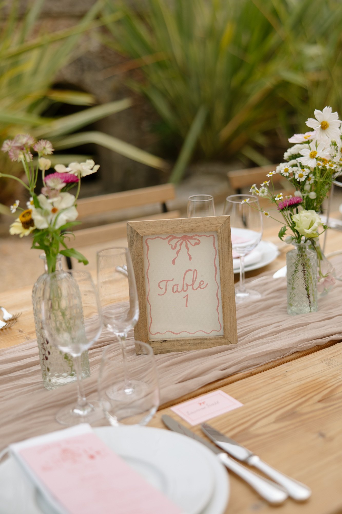 A wooden table set for an event with plates, glasses, flower arrangements in glass vases, and a framed sign reading Table 1 in the center.