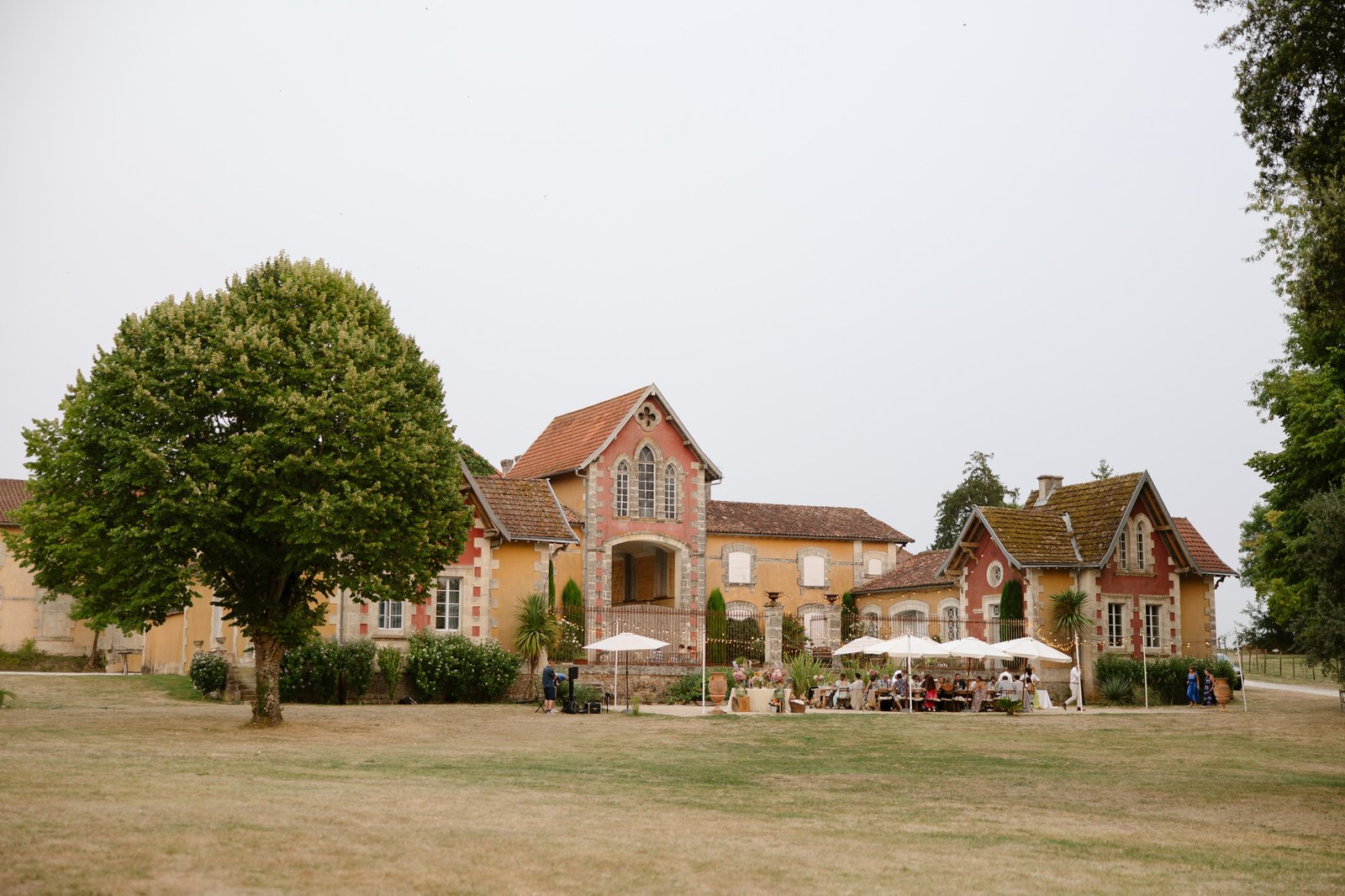 Large country house with rustic yellow and red exteriors, surrounded by a lawn and trees; people sit at tables with umbrellas in front of the building. Captured by Cognac wedding photographer.