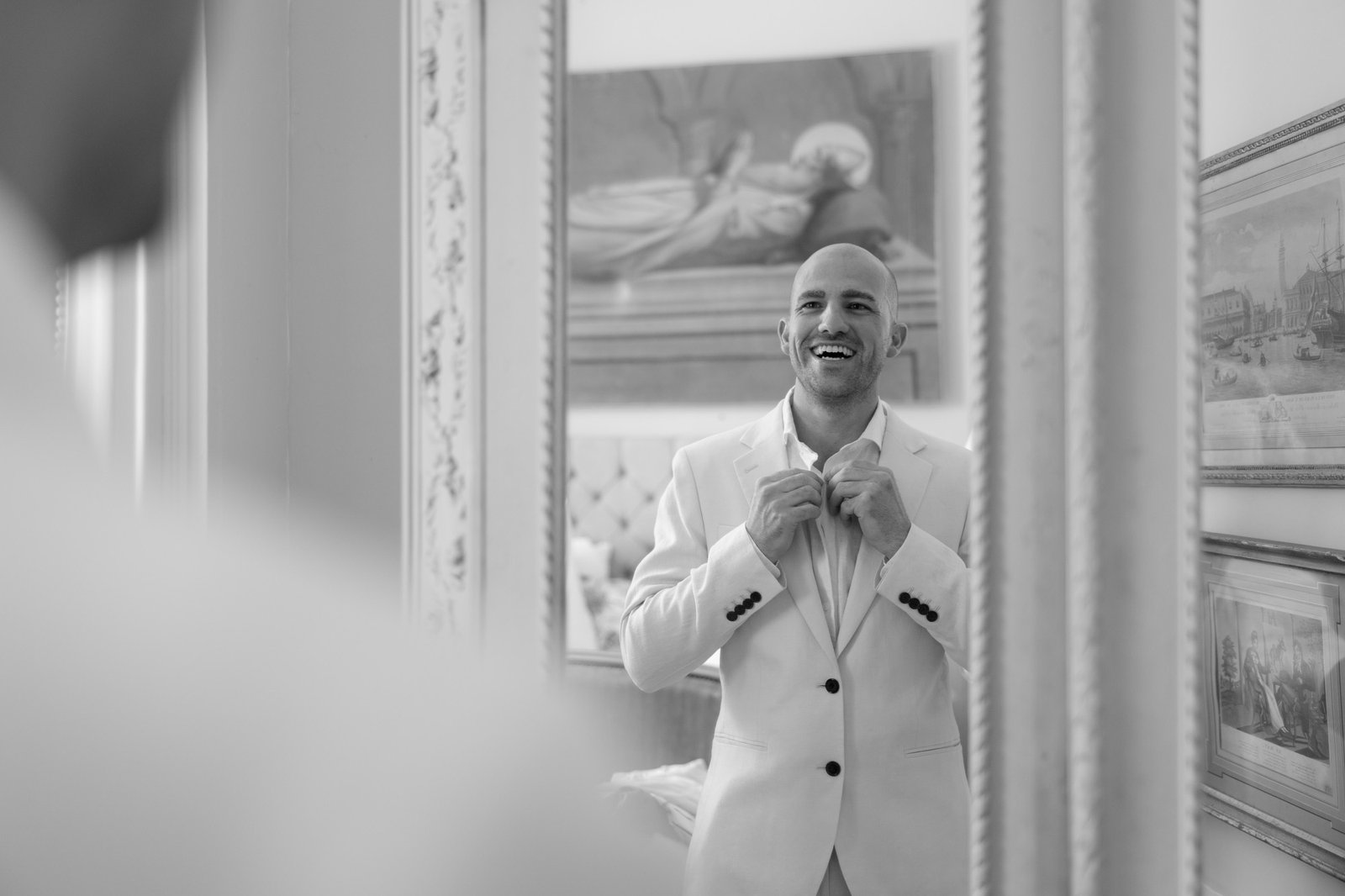 A man in a white suit adjusts his collar and smiles at his reflection in a mirror, with framed artwork visible on the wall behind him.