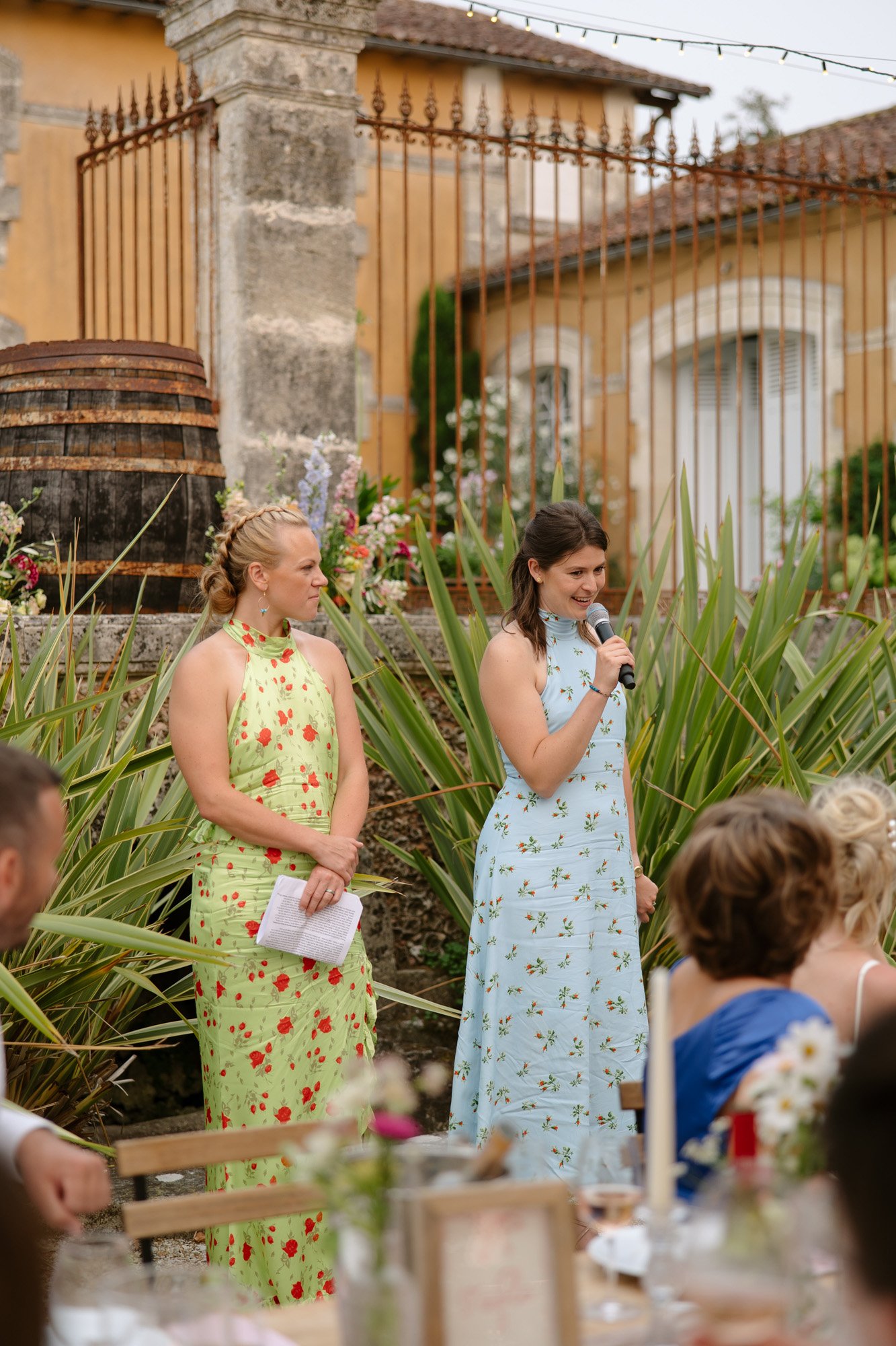 Two women in floral dresses stand outdoors; one holds a microphone, the other holds papers, speaking to seated guests at an event.