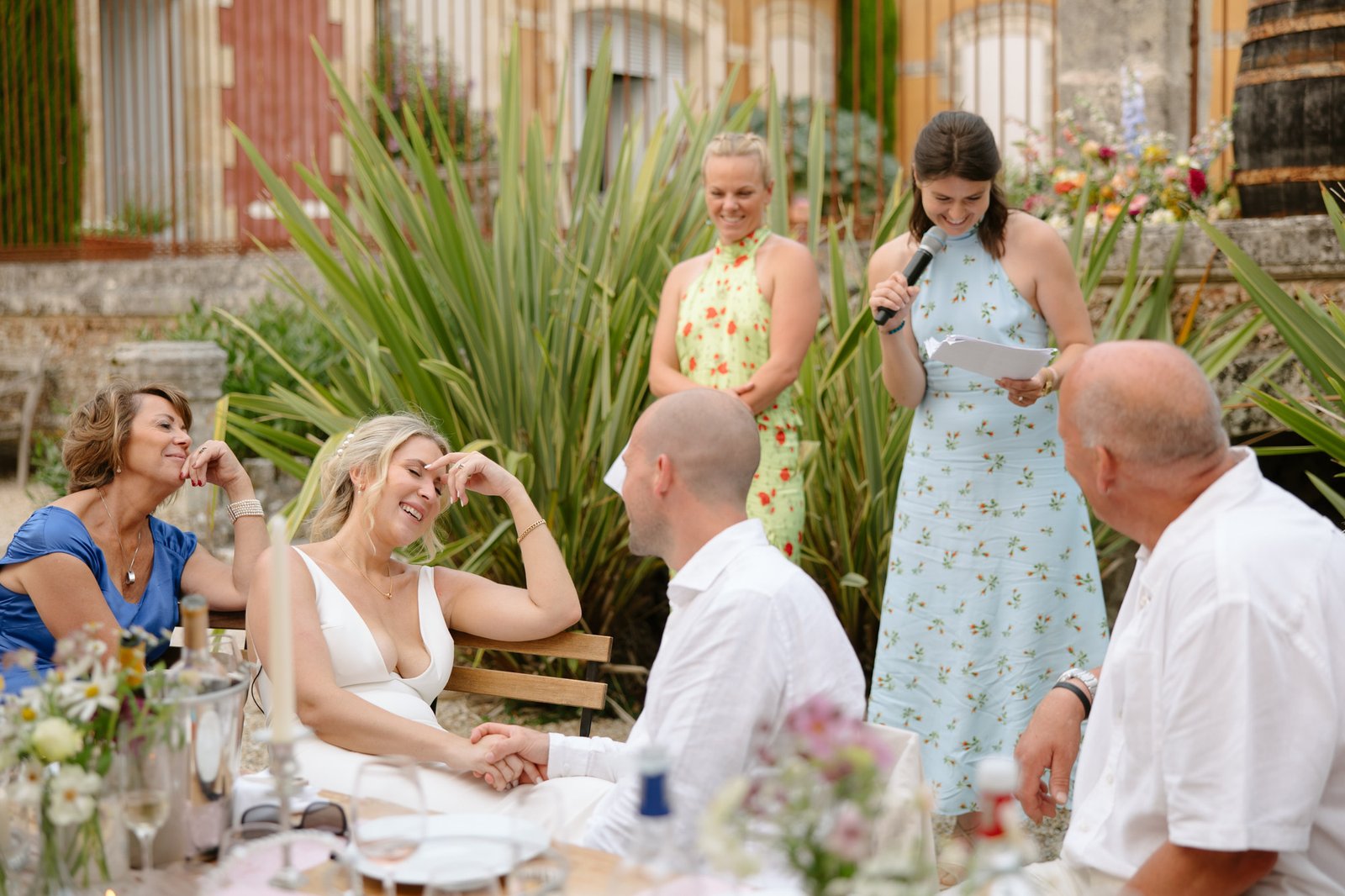 A woman in a blue dress reads from a microphone to a seated couple, who are holding hands and laughing, during an outdoor gathering with others present.