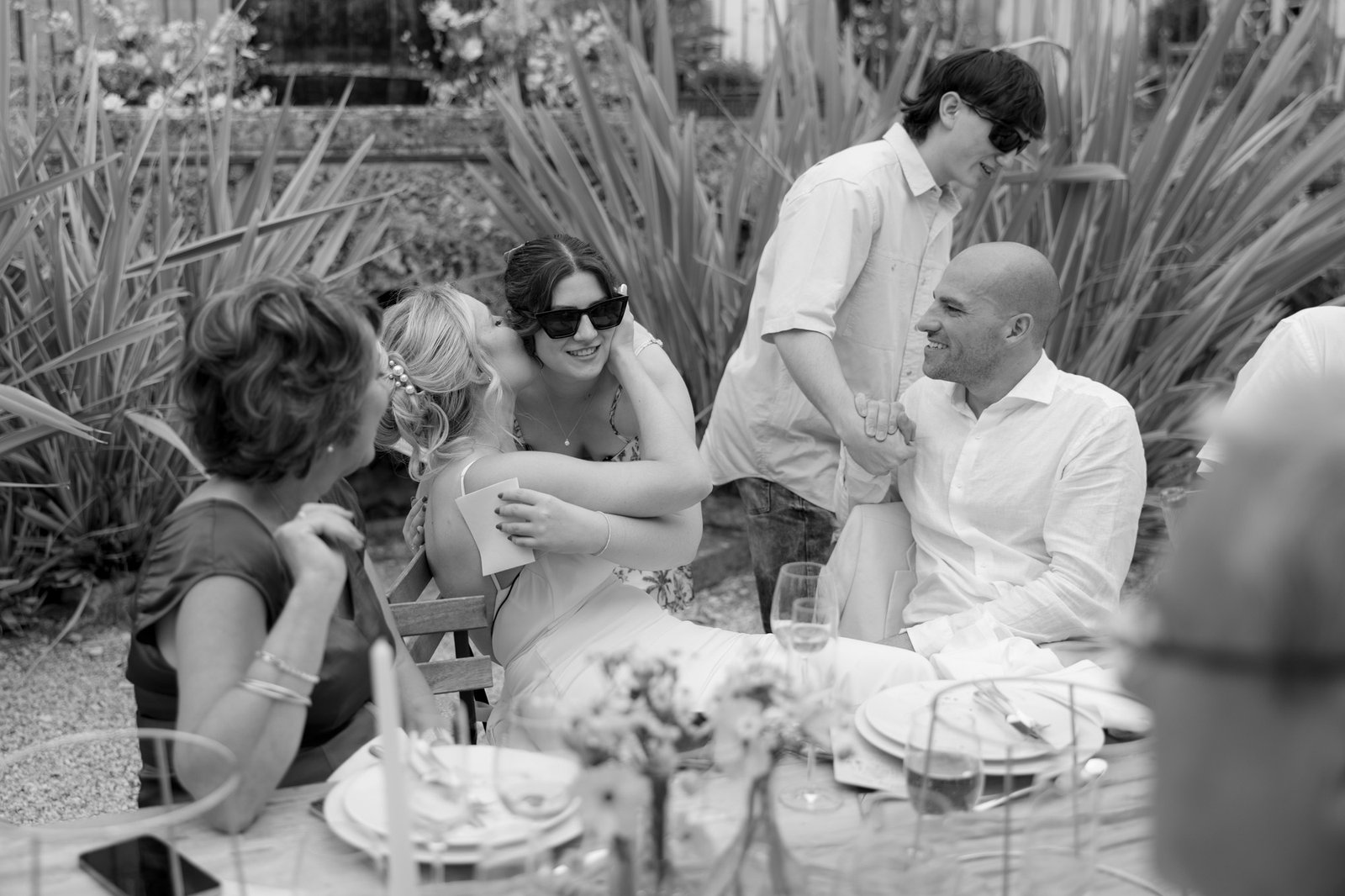 A group of people sits at an outdoor table, with two women hugging and others smiling and interacting in a garden setting.