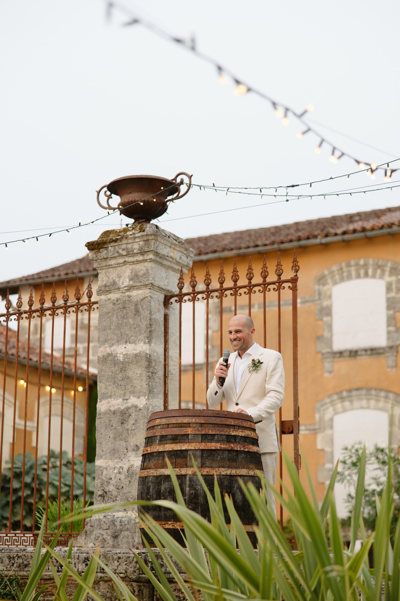 A man in a beige suit stands behind a wooden barrel, speaking into a microphone outdoors near a stone pillar and iron fence, with a rustic building in the background.