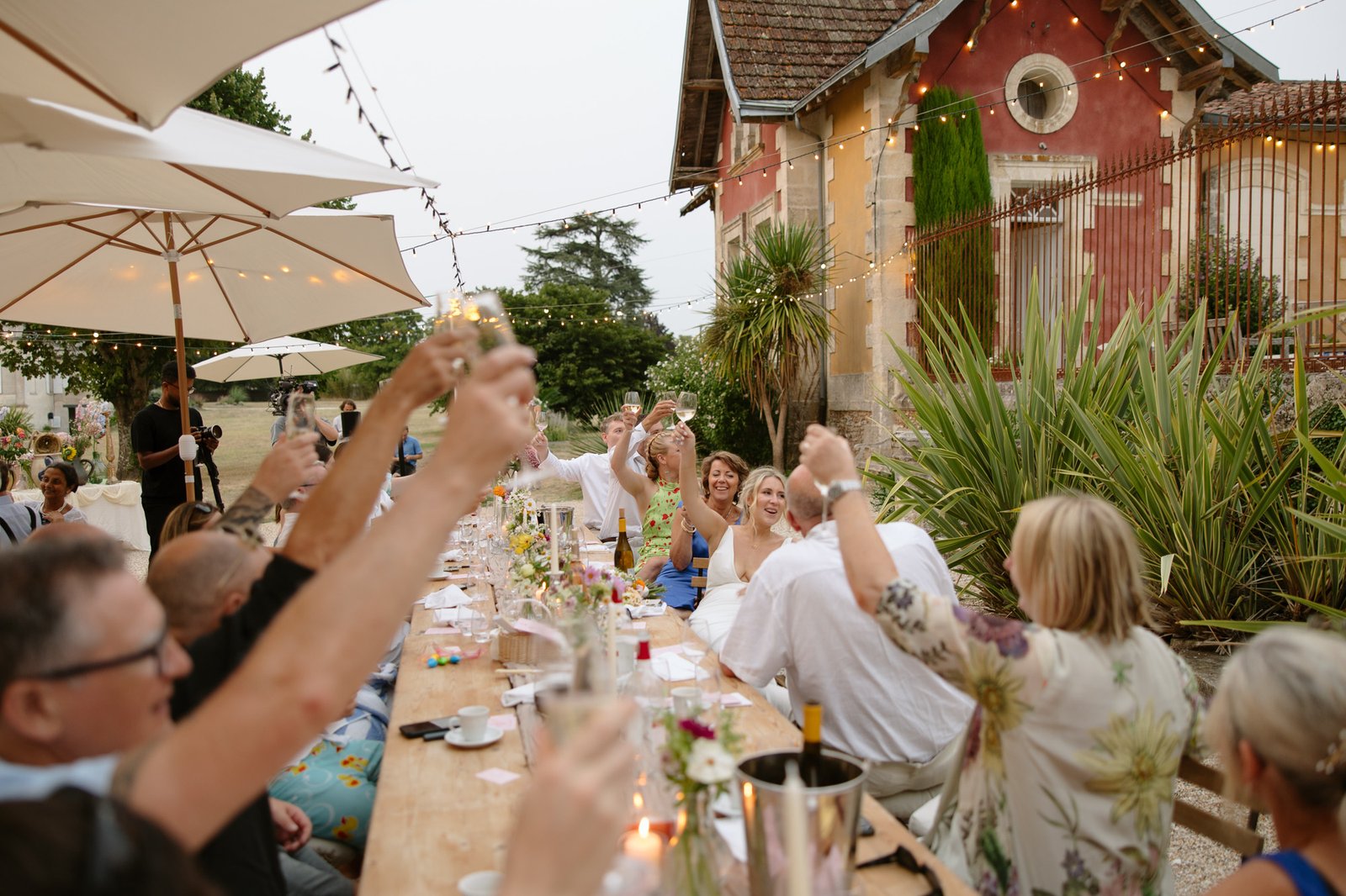 A group of people seated at a long outdoor table raise their glasses in a toast, with string lights and a rustic building in the background.