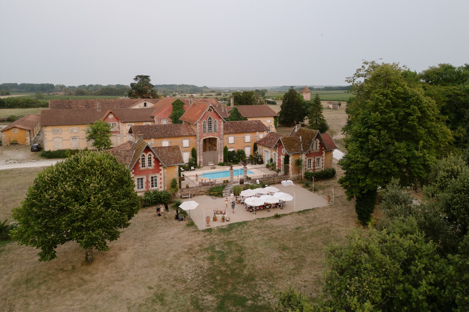 Aerial view of a rustic estate with red-roofed buildings, a central pool, and an outdoor dining area set with tables and umbrellas, surrounded by trees and fields.