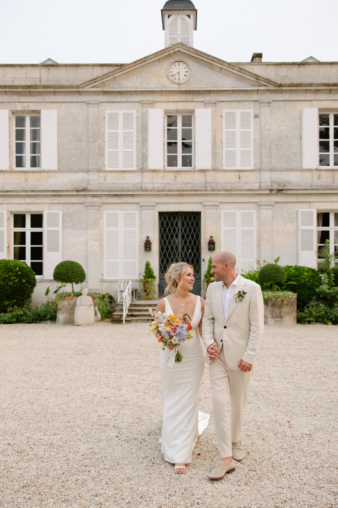 A bride and groom in white attire walk hand-in-hand outside a large, elegant building with white shutters and a clock above the entrance.