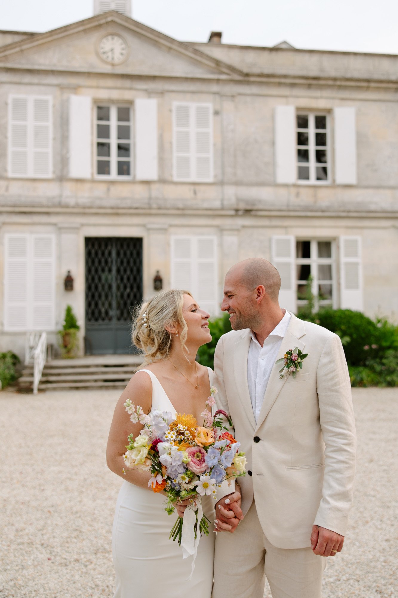 A bride and groom stand smiling and holding hands in front of a large, light-colored building with shuttered windows. The bride holds a colorful bouquet.