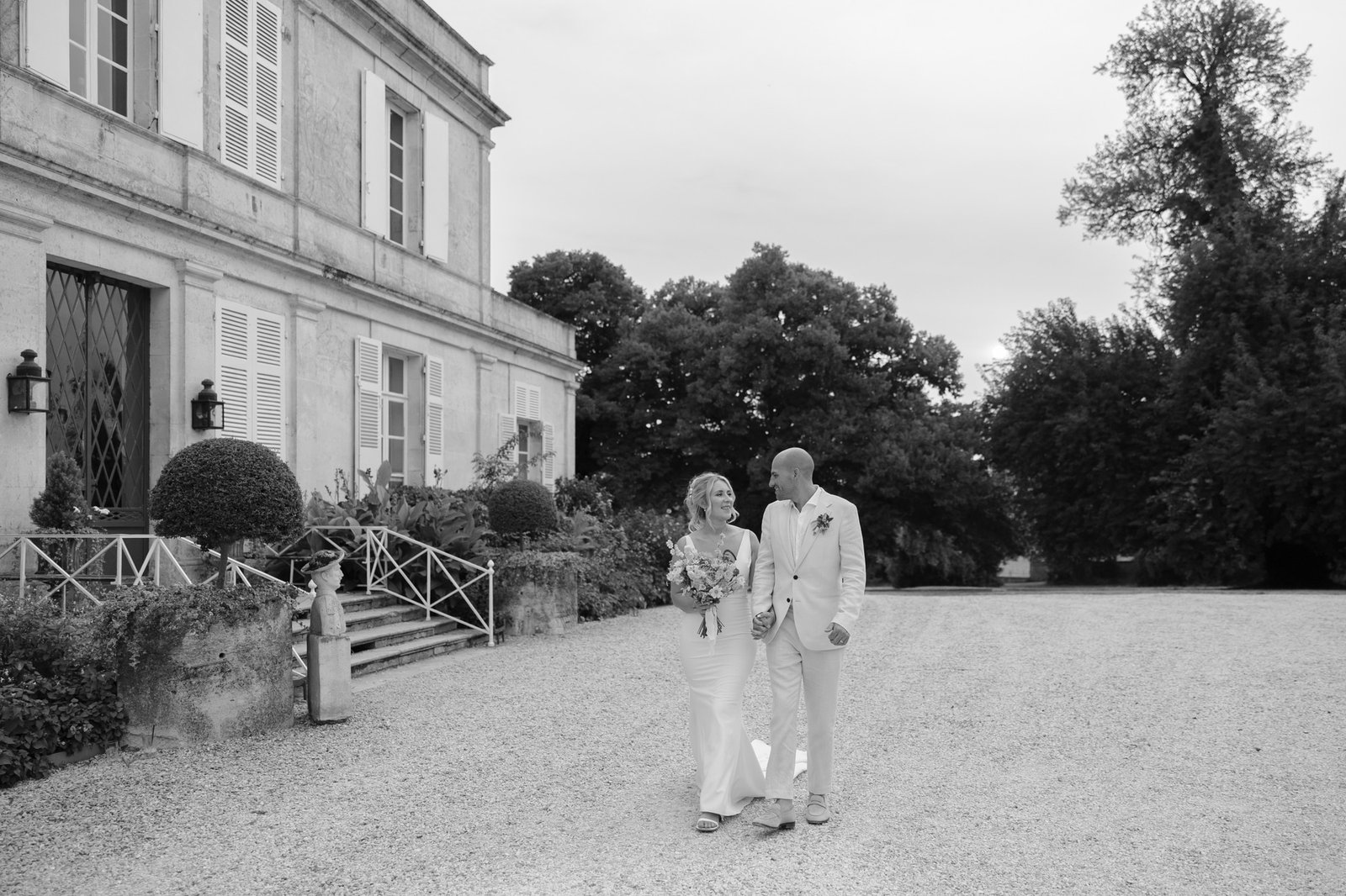 A bride and groom in formal attire walk hand in hand outside a large building with trees and gravel surroundings. Captured by Cognac wedding photographer.