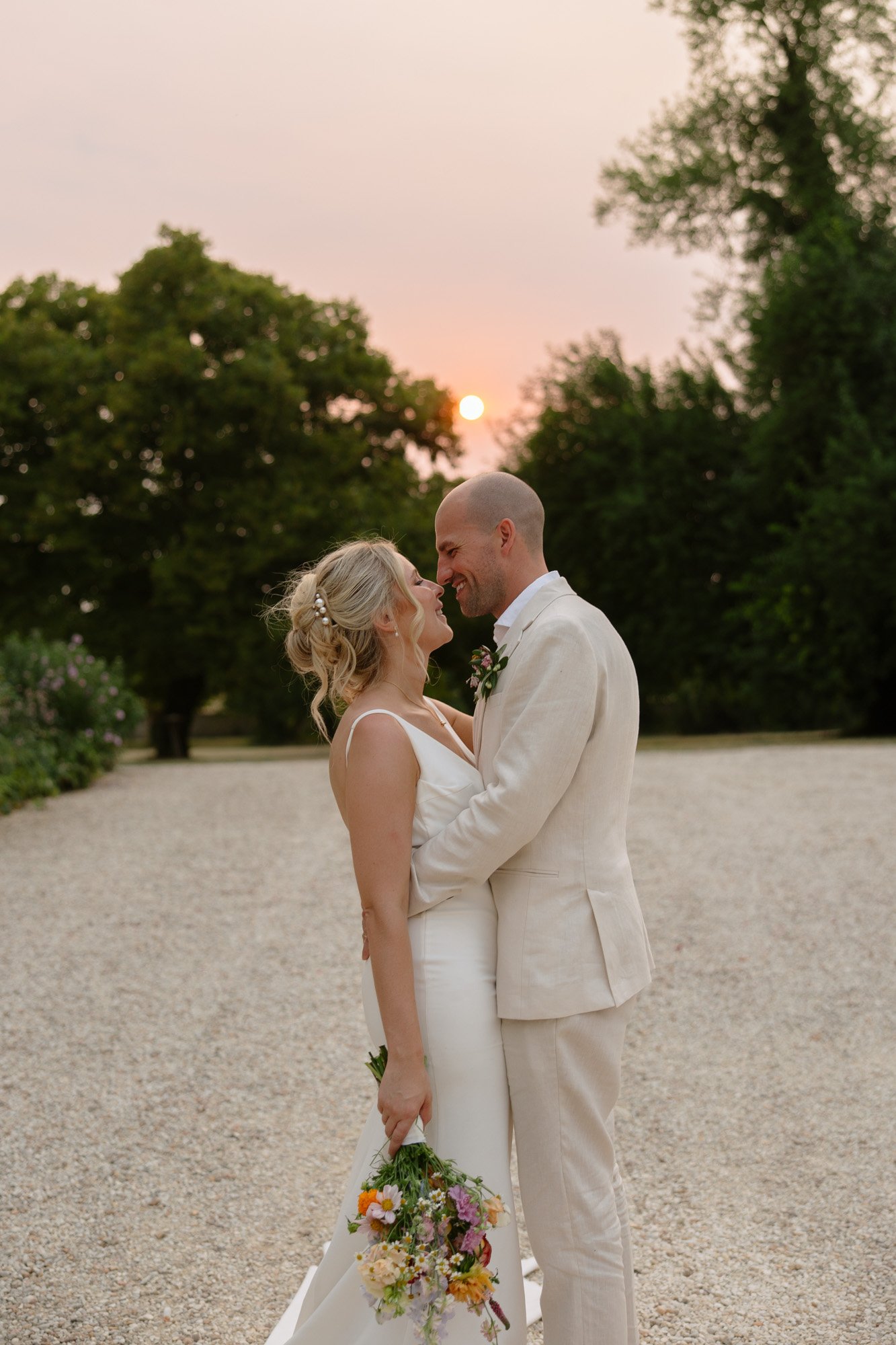 A bride and groom in white attire stand close together, smiling at each other, with the bride holding a bouquet and the sun setting behind them.