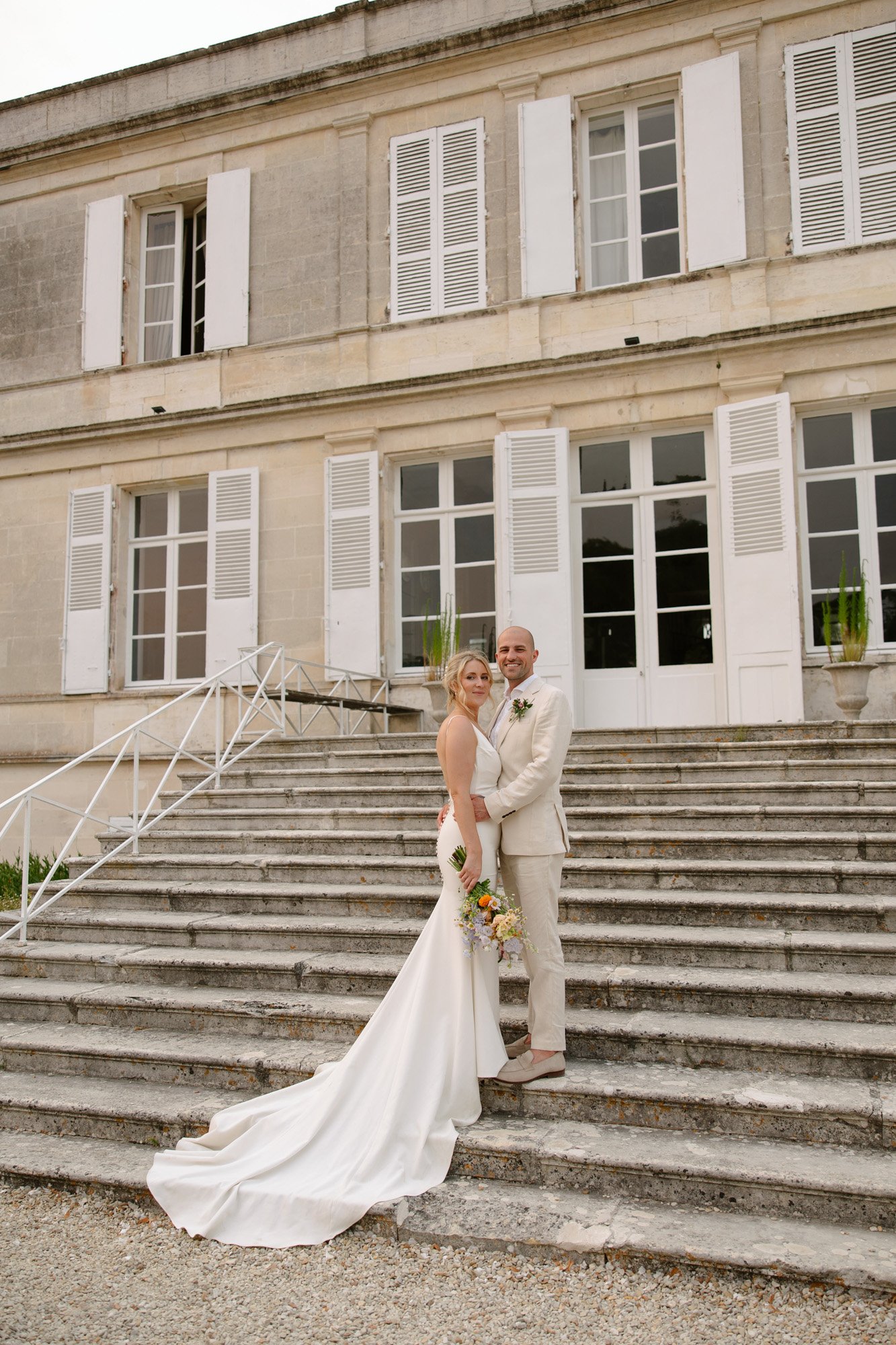 A bride and groom in formal wedding attire stand on stone steps in front of a large building with white shutters.  Captured by Cognac wedding photographer.