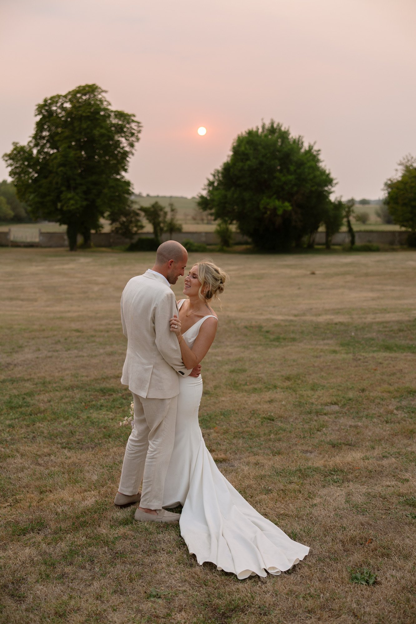 A bride and groom in formal wedding attire stand closely together outdoors on a grassy field at sunset, with trees and a low fence in the background.