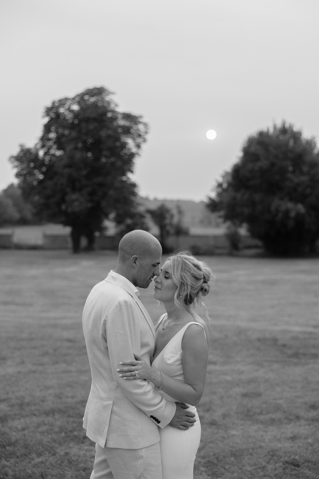A couple in formal attire stands closely together outdoors on grass, facing each other with eyes closed, with trees and the sun in the background.
