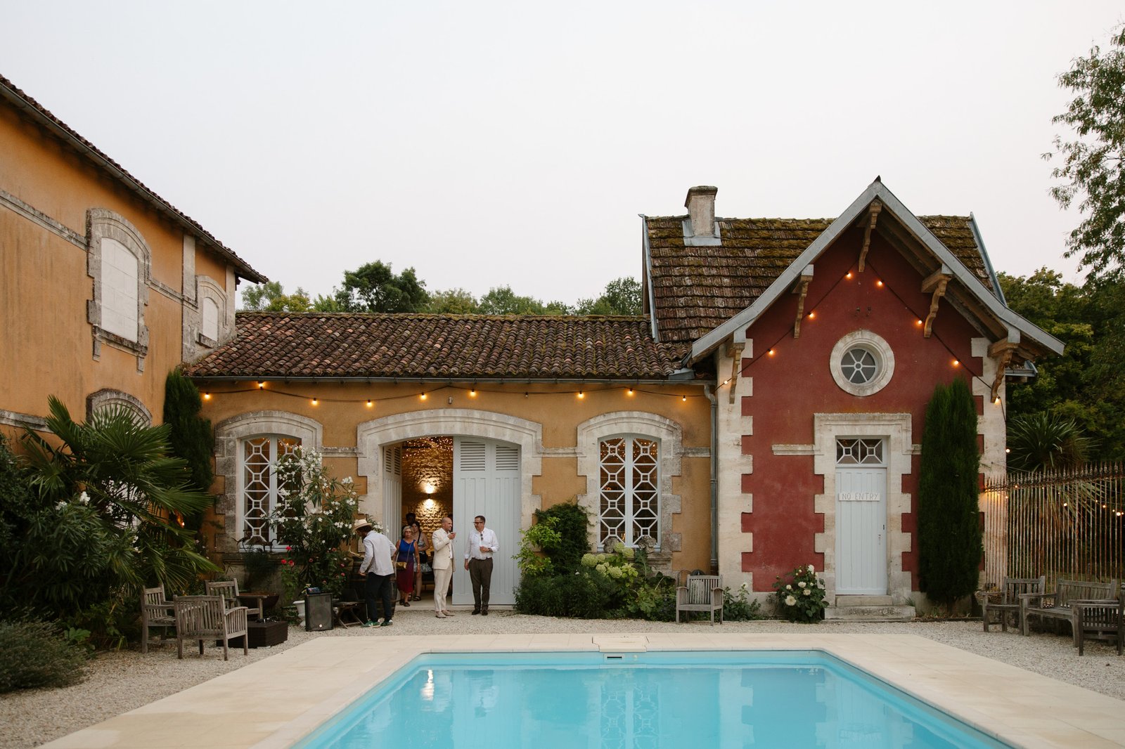 A small group of people stand by the entrance of a rustic house with string lights, next to a swimming pool and surrounded by greenery.