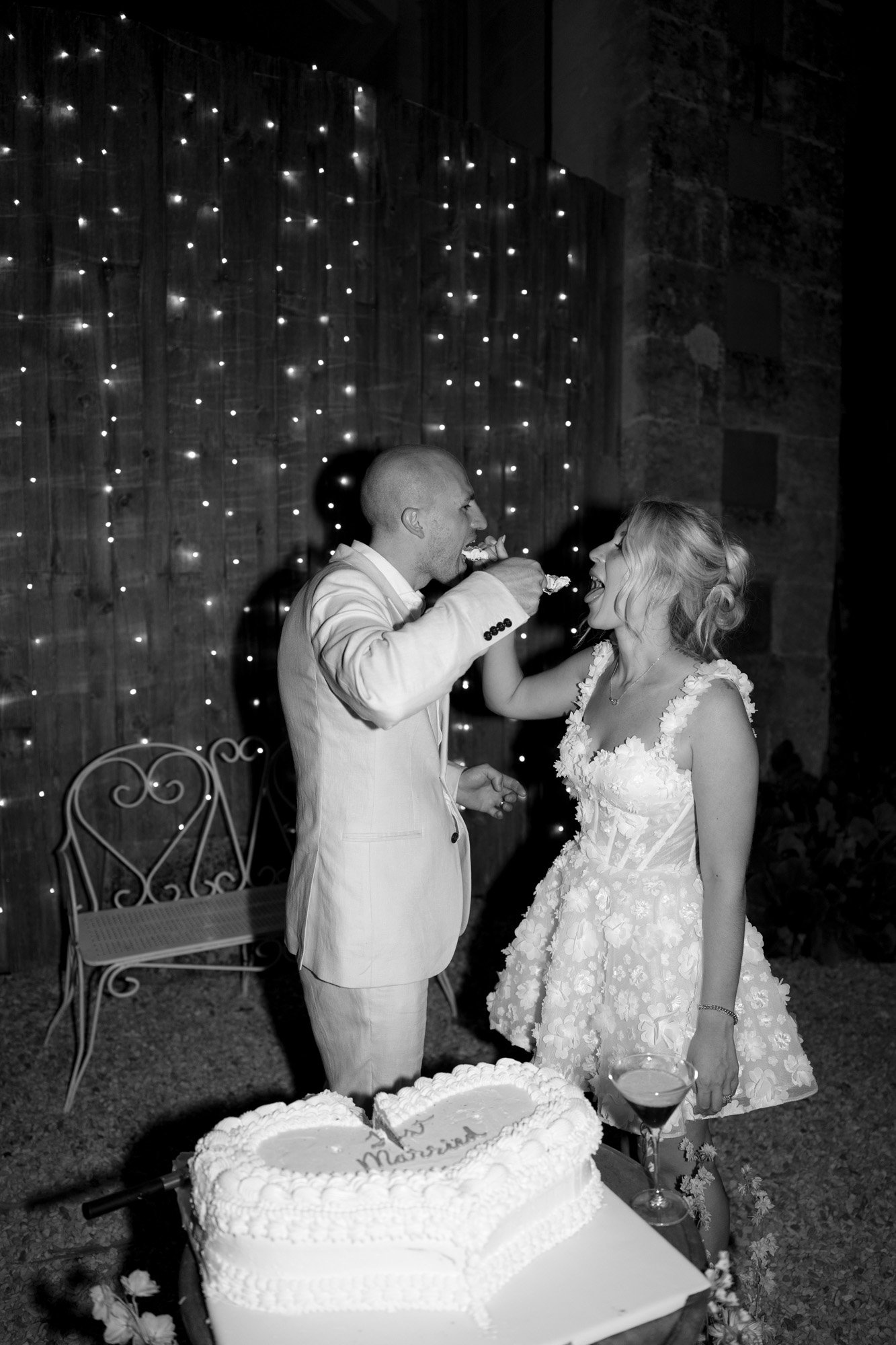 A couple in formal attire feed each other cake at night in front of string lights and a rustic wall; a heart-shaped cake sits on a table nearby.