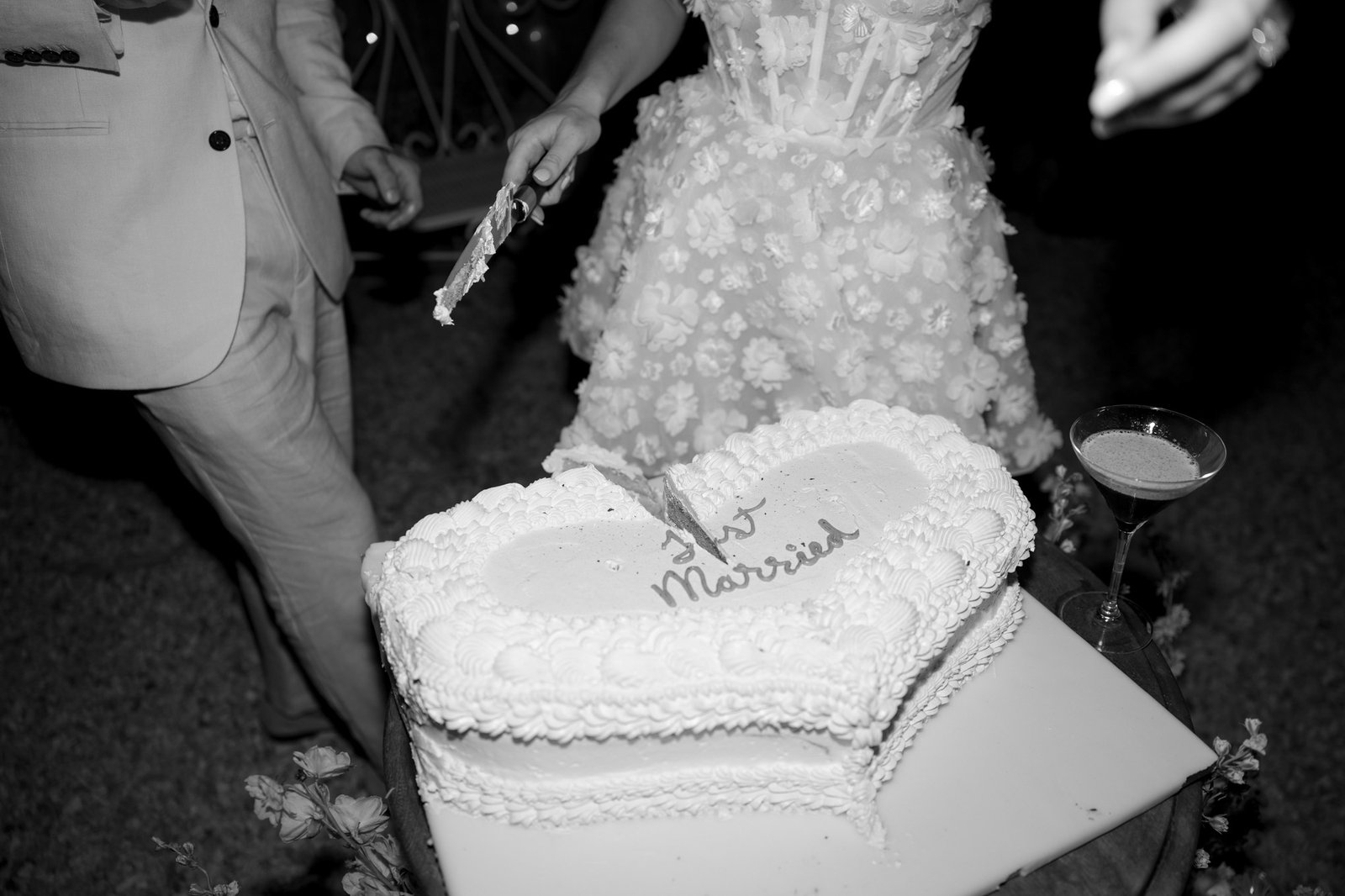 A couple cuts a heart-shaped wedding cake with Just Married written on it; only their torsos and hands are visible.