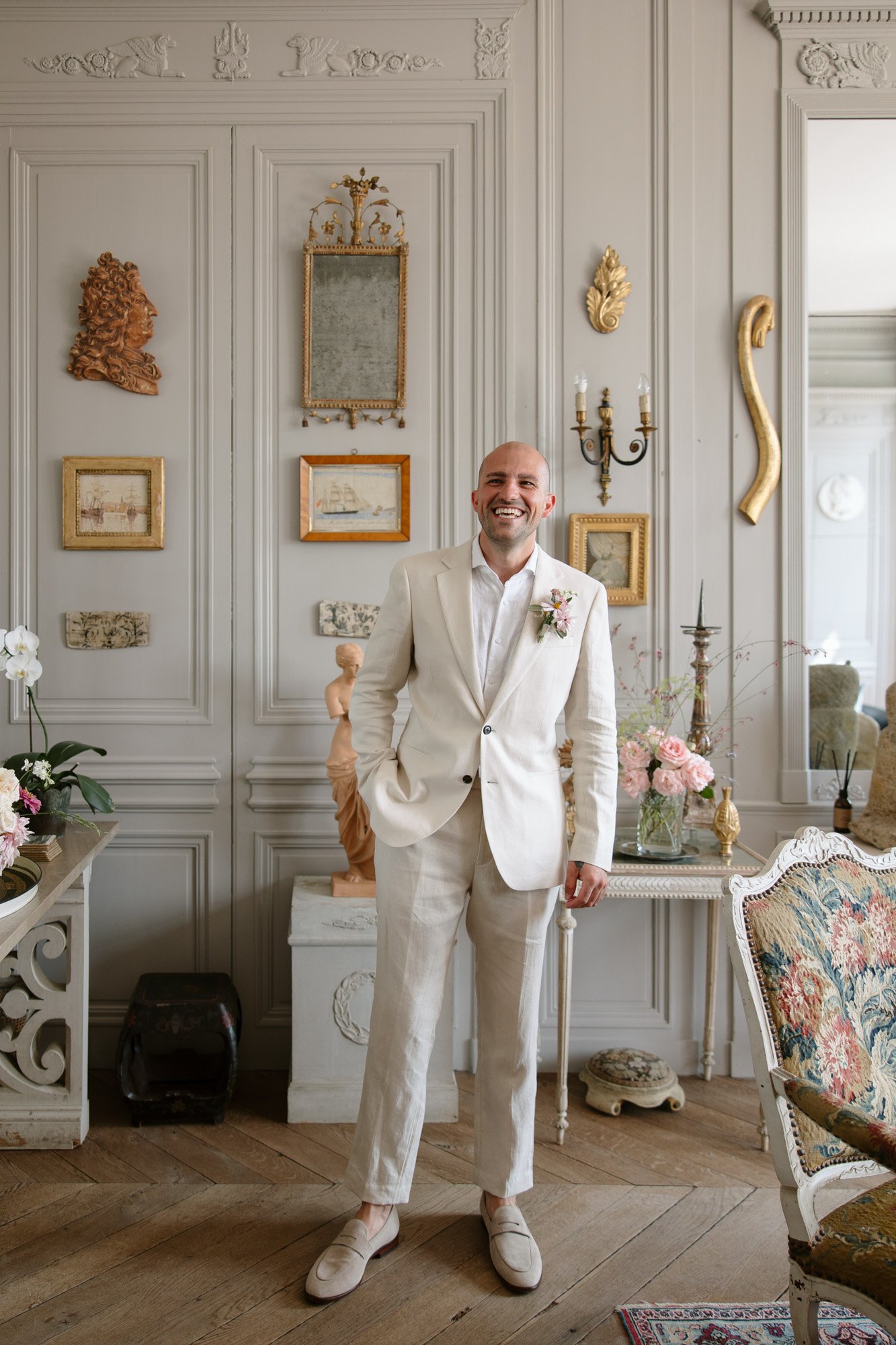 A man in a white suit stands smiling in an ornately decorated room with classic wall moldings, art, flowers, and antique furniture.