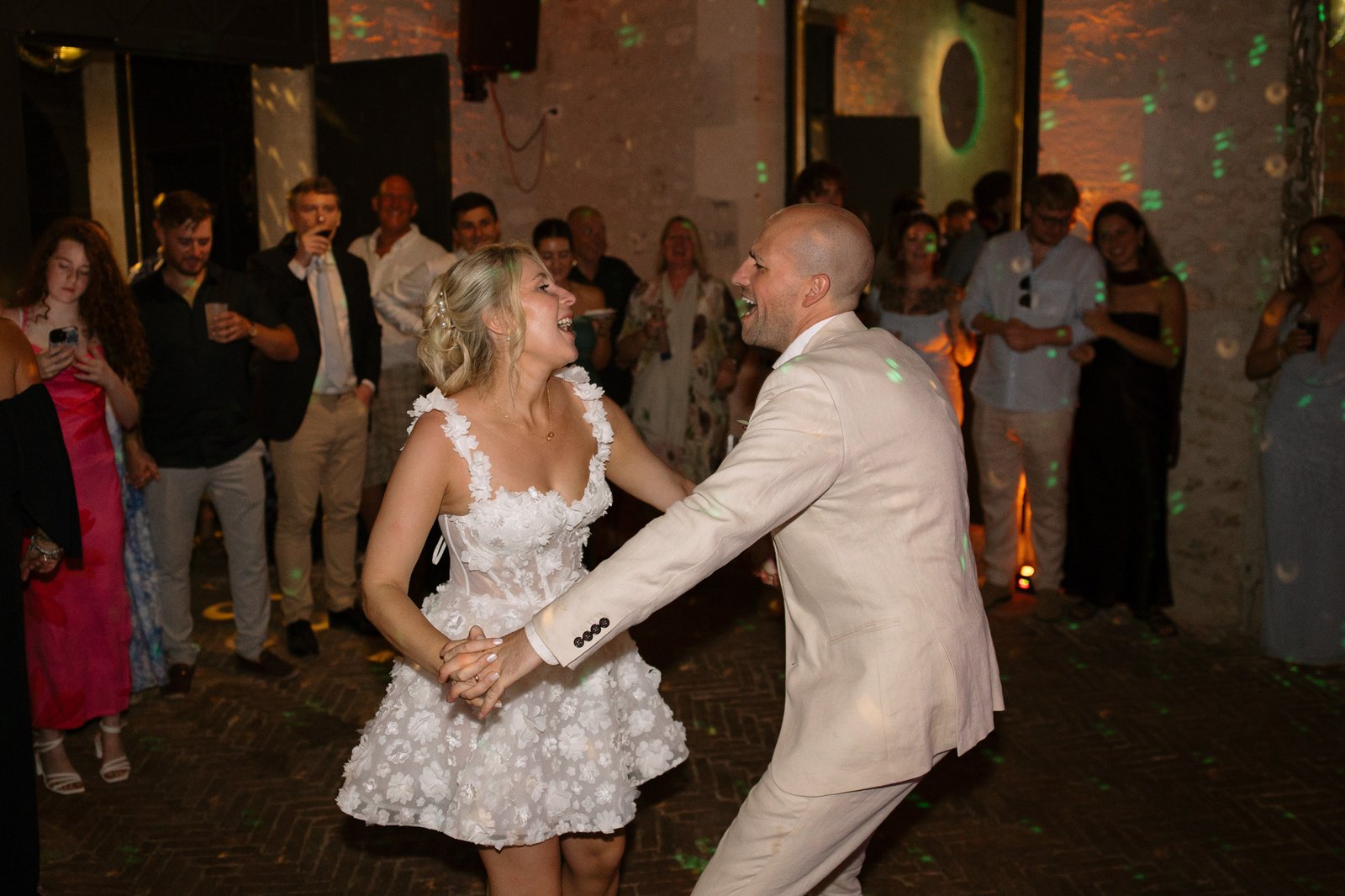 A bride and groom dance together at their wedding reception while guests watch and smile in the background.