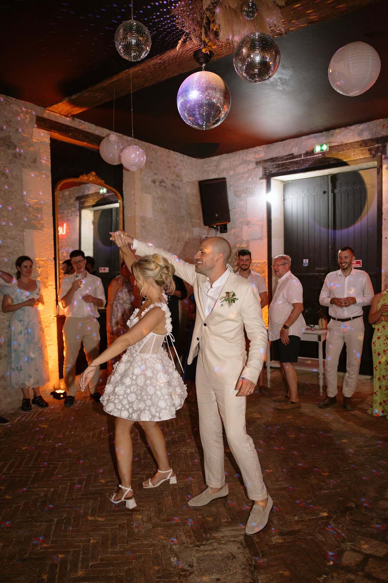 A bride and groom dance together at their wedding reception in a dimly lit venue with disco balls and guests watching in the background.